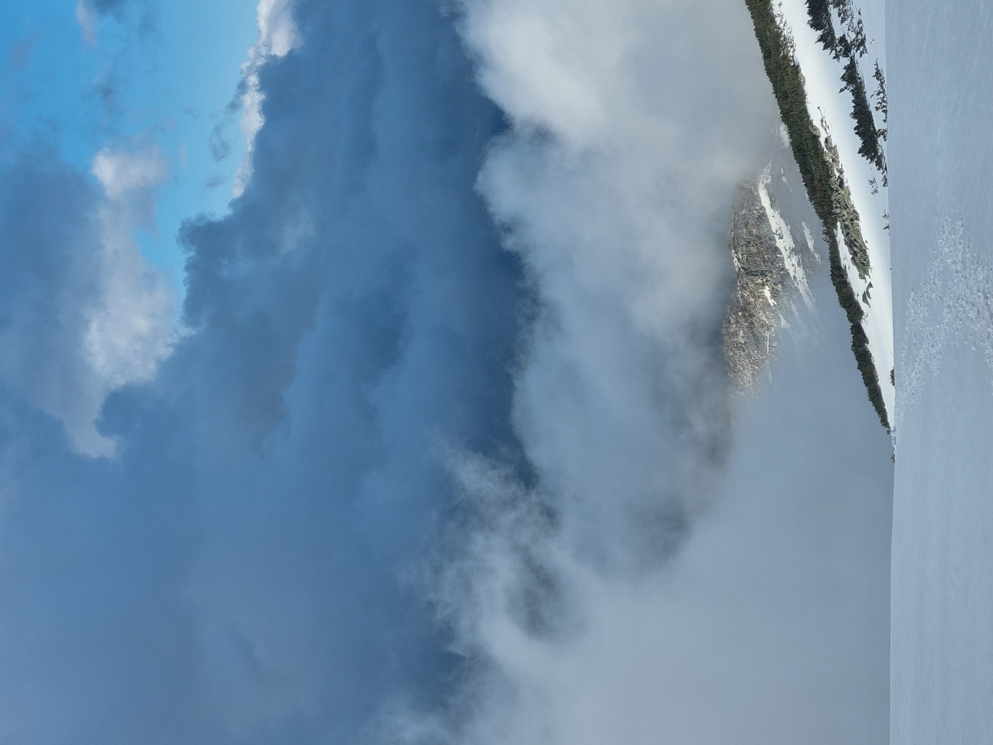 An outdoor landscape scene with clouds obscuring the upper left portion of the image. The clouds transition from darker, more opaque masses to thinner, wispy formations. The right side of the image depicts a snow-covered mountainous terrain. Sparse vegetation, possibly coniferous trees, is visible along the upper edge of the snow line. Some areas on the mountain appear rocky. The landscape is potentially located in the region of Govedartsi, Bulgaria. The image appears to be rotated clockwise, placing the sky on the left side.