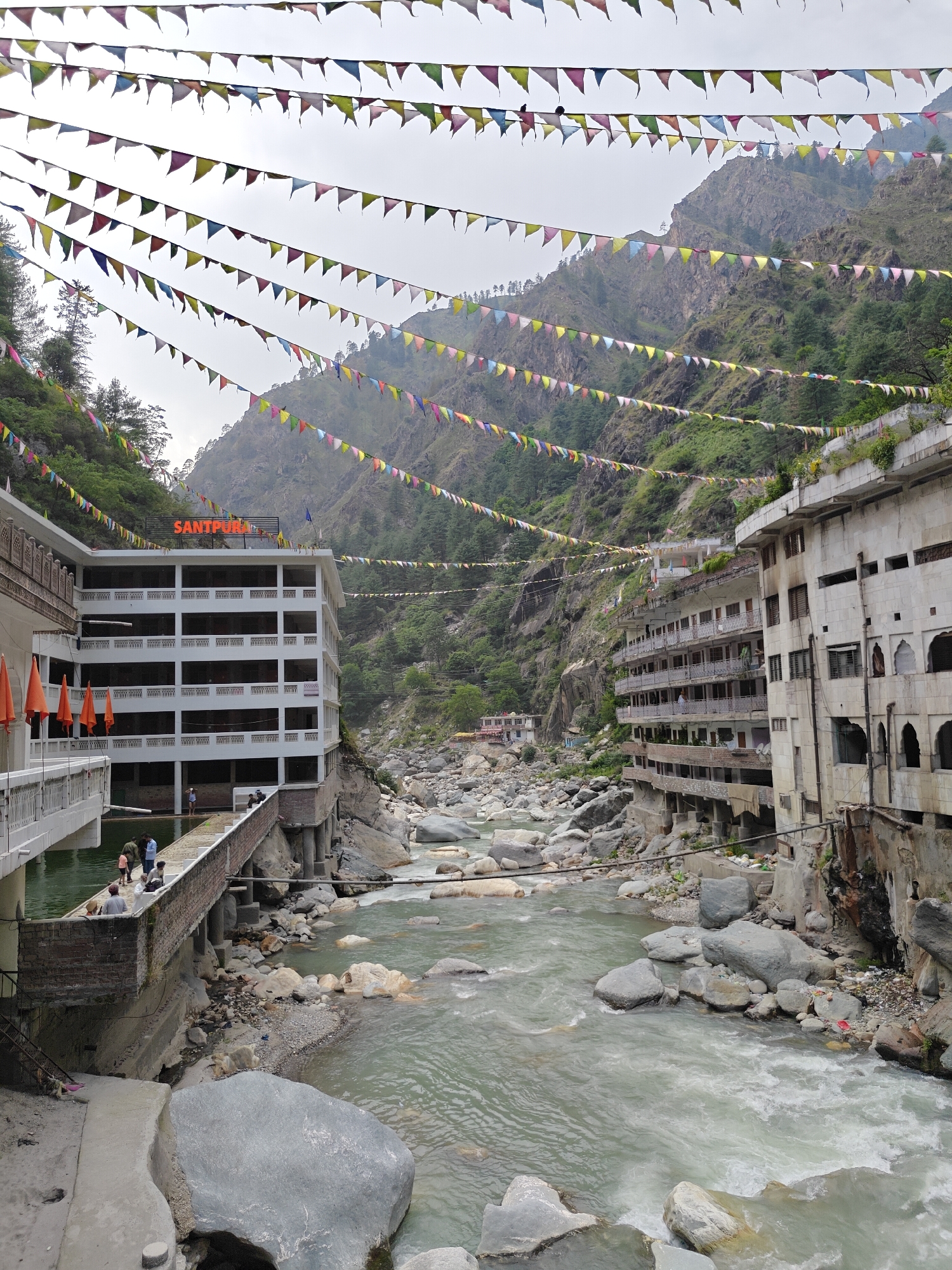 The image captures a vibrant and rugged mountain valley in Kasol, India, under an overcast sky. Numerous colorful triangular flags, strung across the valley, dominate the upper and mid-ground, creating a festive atmosphere.
A turbulent river, characterized by its grey-green water and numerous large, smooth boulders, flows through the center of the frame, originating from a distant, mist-shrouded gorge. On the left bank, a multi-story white building with multiple balconies stands prominently. Its rooftop features an orange sign with visible text "SANTPURA." Below this sign, several orange flags are displayed. People are visible on a lower walkway and a bridge-like structure extending from this building, some gathered in small groups, observing the river or socializing. The building itself has uniform rows of windows and balconies with intricate white railings.
On the right bank, an older, more weathered multi-story building, also with multiple balconies and built into the steep, rocky hillside, contrasts with the white structure. This building appears more rustic, with arched window openings on its lower levels. Patches of green vegetation grow on its upper ledges. The steep mountain slopes, covered with dense green trees and rocky outcrops, rise on both sides of the river, forming the backdrop of the scene. Near the base of the right building, close to the river, some debris and trash are visible. The time of day appears to be daytime, indicated by the overall light, though the sky is uniformly cloudy.