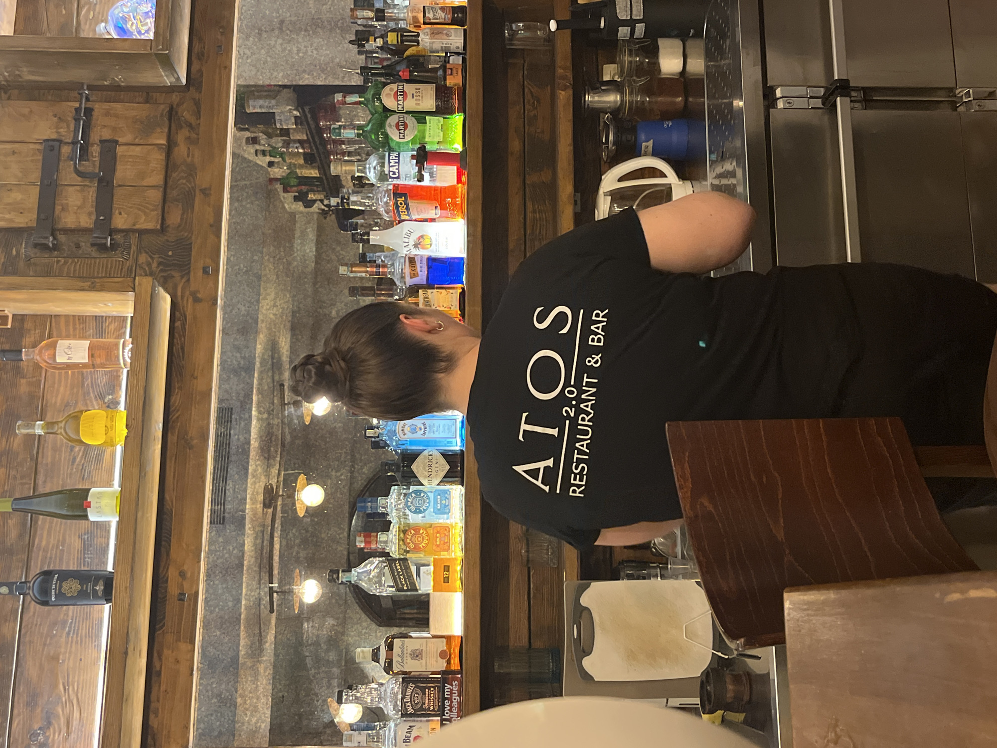 The image captures a scene inside the ATOS 2.0 Restaurant & Bar in Budva, Montenegro. A woman wearing a black T-shirt with the restaurant's logo is visible. Behind her, a bar setup displays an array of alcoholic beverages, including whiskies, vodkas, gins, and liqueurs. The bar shelves are wooden. A wooden structure with wine bottles is seen on the left side of the image. In front of the woman, a wooden surface and a cutting board are visible.