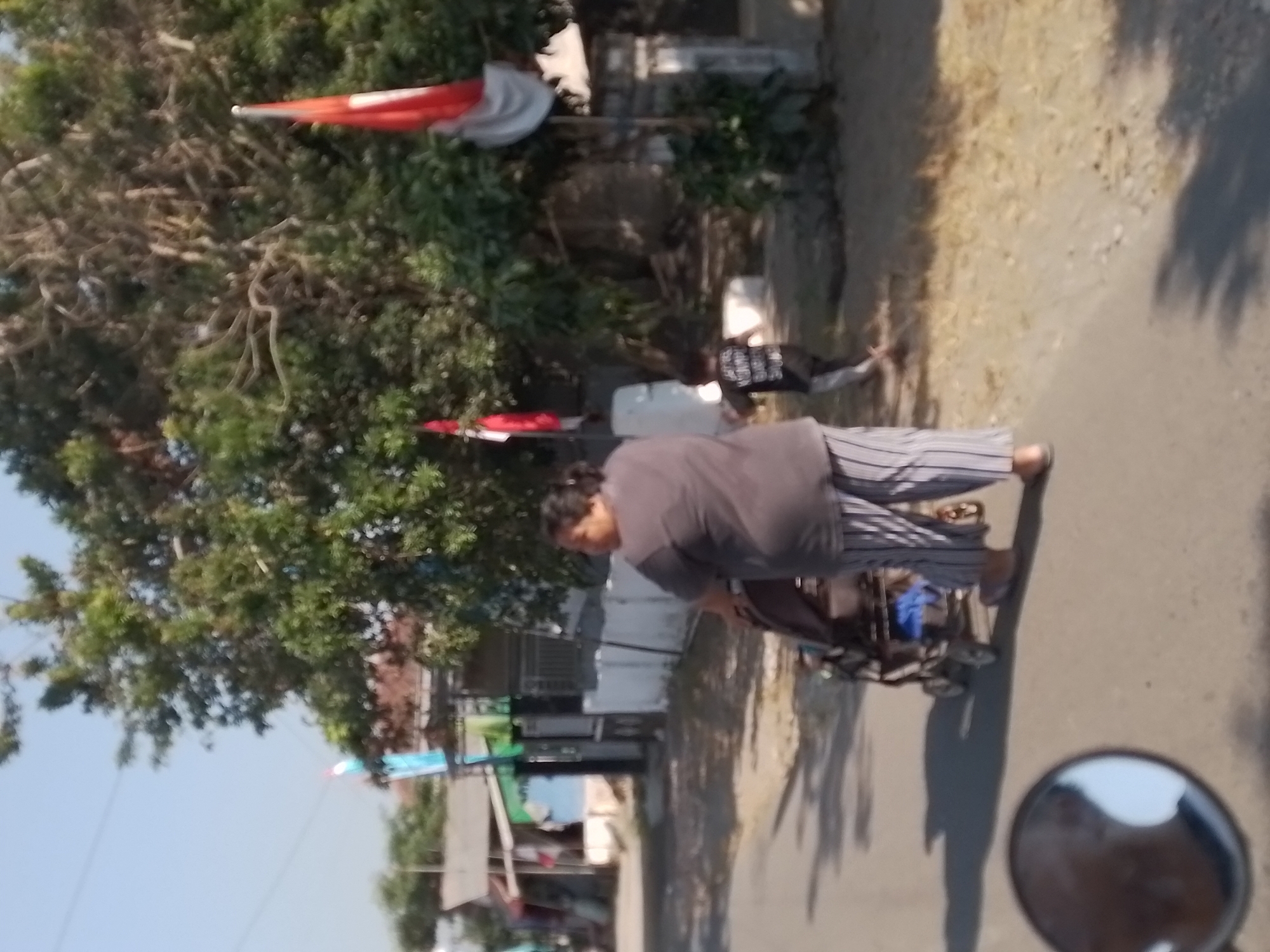 In Wates, Indonesia, on a paved road with dry grass and dirt on the side, a woman is pushing a stroller. She is wearing a dark grey t-shirt and grey and white striped pants. A young child in a black t-shirt with white text is running away from the woman and stroller, towards a large tree. Behind the tree, two red and white Indonesian flags are visible, one on a pole and another attached to a building. In the foreground, a motorcycle's side mirror reflects a person's face. Buildings are visible in the background.