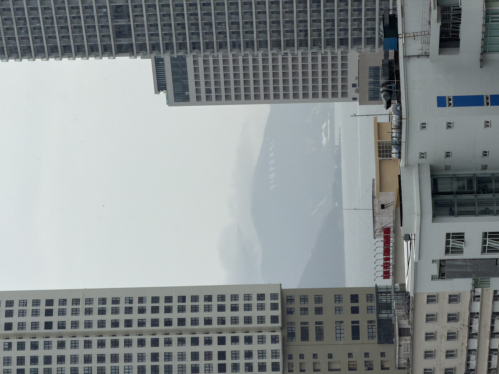 The image presents an urban scene in Nha Trang Ward, Vietnam. Several high-rise buildings with repetitive window patterns frame the view. Between the buildings, a hazy mountain range is visible with the word "Vinpearl" displayed on its side. To the right, the side of a white building with a red stripe along the edge and assorted structures.