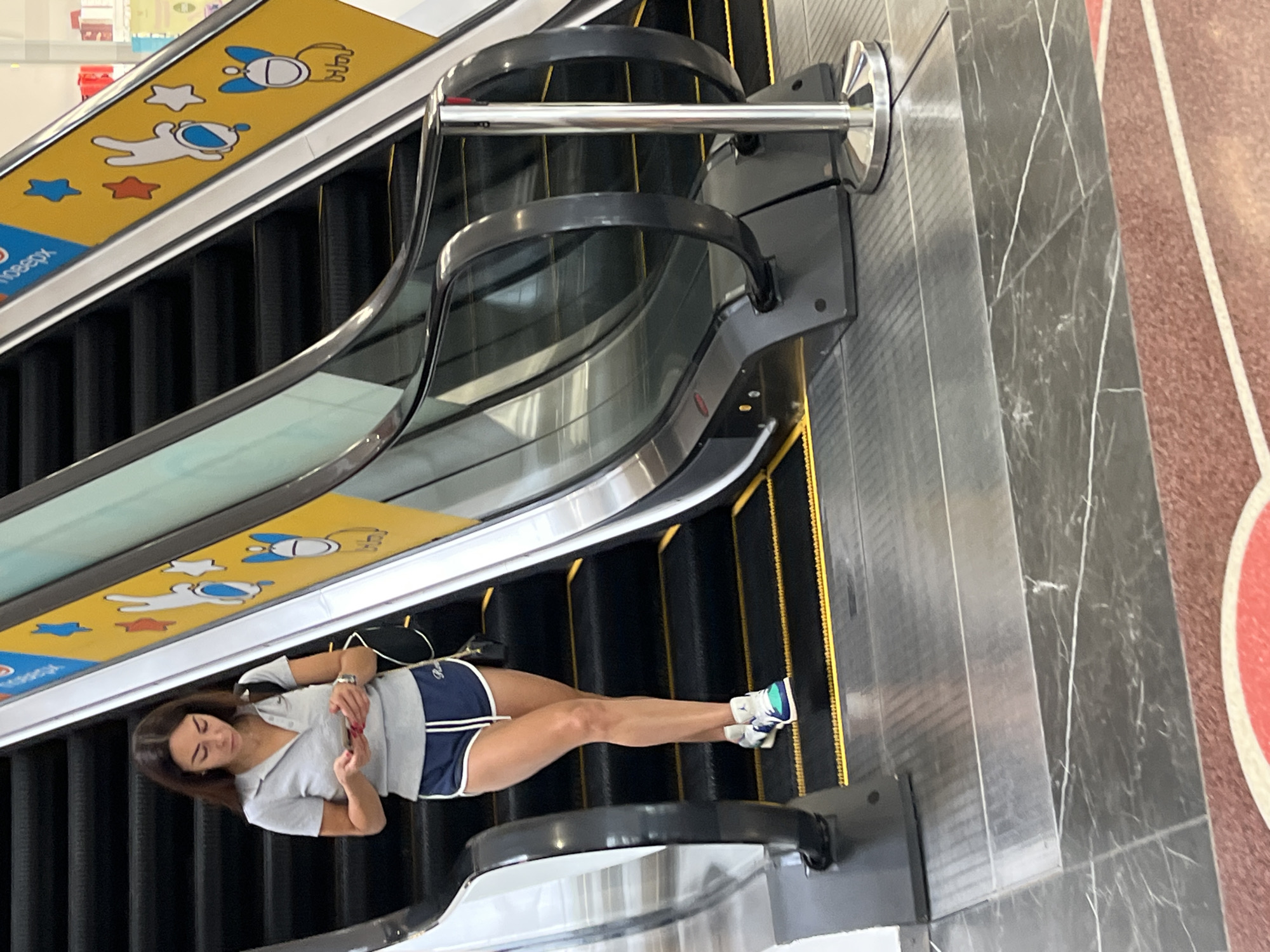 A woman is standing on an escalator, looking at her phone. She is wearing a grey polo shirt, navy blue shorts with white trim, and athletic shoes. The escalator has yellow warning stripes and a yellow decorative panel with cartoon characters. The woman is holding her phone in her right hand and her left hand is resting on the escalator railing. The floor is tiled with a marble pattern. The image is taken from a slightly elevated angle. The scene appears to be in a public indoor space, possibly a shopping mall. The provided image context specifies the location as Kharkiv, Ukraine.