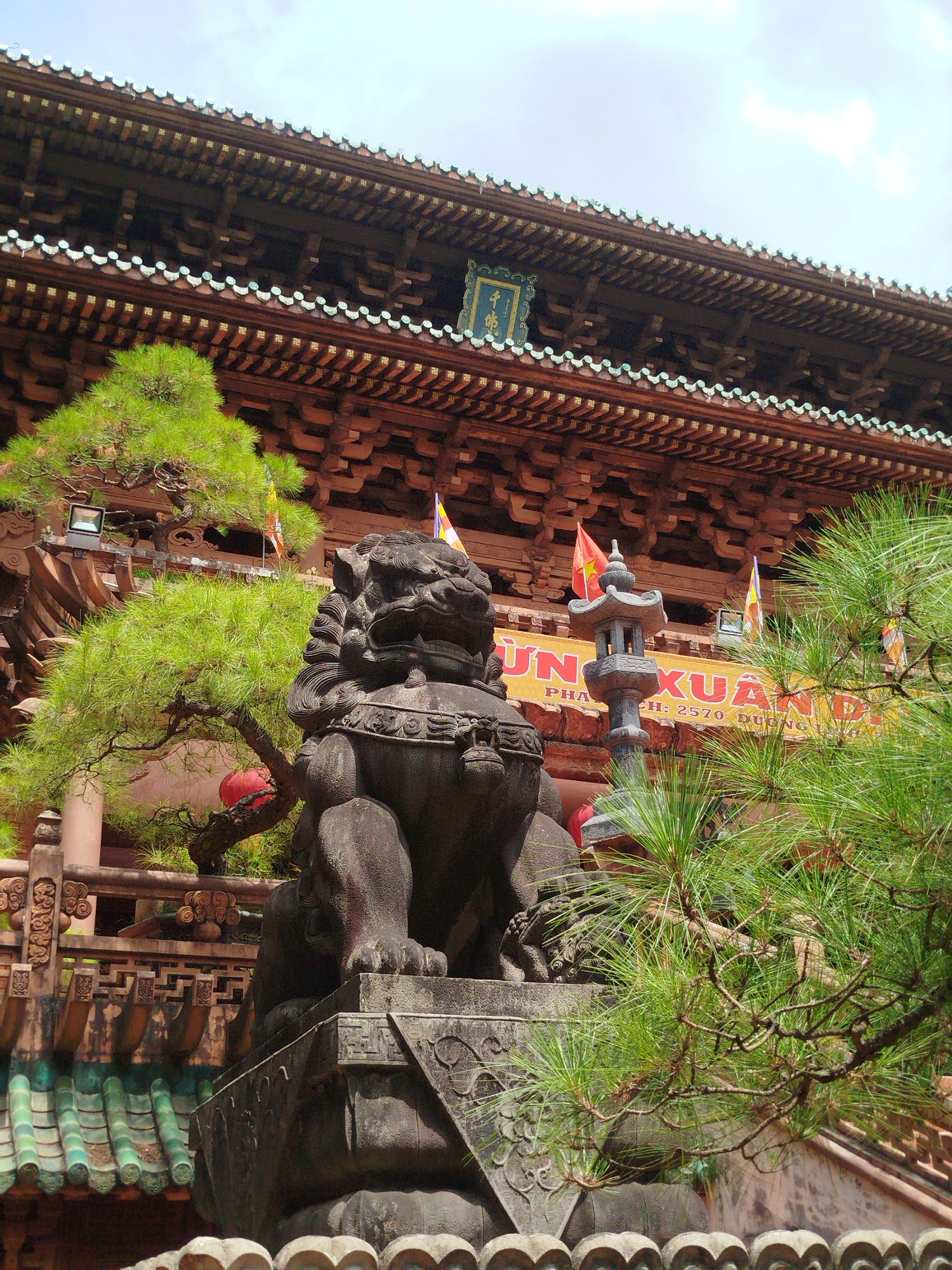 The image depicts a stone lion statue in Hoi Phu Ward, Vietnam. The statue is positioned in front of a Buddhist temple. The temple has an ornate wooden facade with multiple tiers and a sign with gold lettering on a blue background. The statue is surrounded by greenery including a large pine tree and a smaller shrub. Several colorful flags are visible in the background. The sky is partly cloudy.