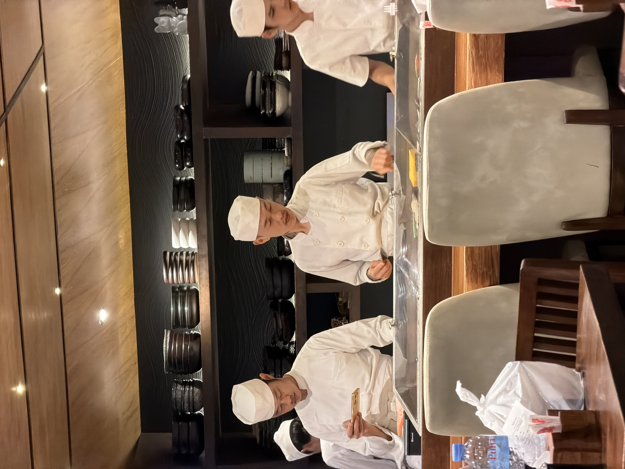 The image depicts a sushi preparation area in a restaurant in San Sai Luang, Thailand. Three chefs, wearing white uniforms and hats, are working behind a clear counter. They are engaged in preparing sushi, handling food items and tools. Behind the chefs are decorative shelves displaying pottery and other items. To the right of the counter are chairs with light-colored upholstery and dark wood frames. A plastic bag and a bottle of water are visible near the chairs. The restaurant's decor includes wood paneling and textured wall designs.