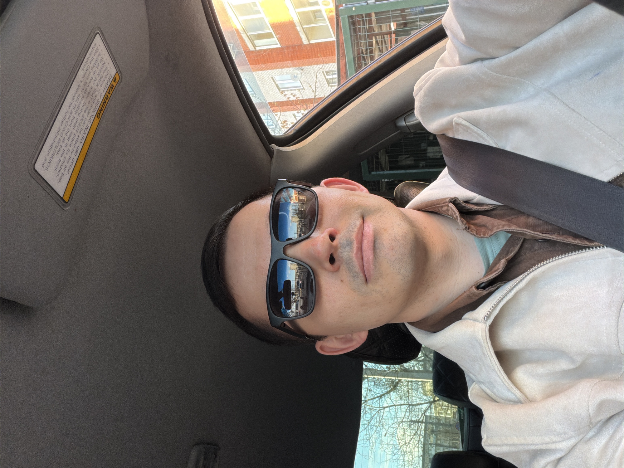 The image shows a man taking a selfie inside a car, rotated 90 degrees counter-clockwise. He is seated in what appears to be the driver's seat, wearing dark sunglasses, a light-colored jacket over other layers, and a dark seatbelt across his chest. He has dark hair pulled back and light facial stubble, with a neutral to slightly content expression.
The car's interior features a dark headliner and a sun visor on the left, which displays a warning label. A sunroof is visible above, offering a view of a multi-story building with reddish-brown brick walls and numerous windows, brightly illuminated by sunlight. Bare trees are also visible, suggesting an autumn, winter, or early spring season. Reflections in the man's sunglasses also show an urban landscape with buildings and sky. The bright sunlight indicates it is daytime with clear weather.
The warning label on the sun visor reads: "A WARNING DEATH OR SERIOUS INJURY can occur. The BACK SEAT is the SAFEST place for children. Children 12 and under can be killed by the air bag. NEVER put a rear facing child seat in the front. Sit as far back as possible from the air bag. ALWAYS use SEAT BELTS and CHILD RESTRAINTS. See the Owner's Manual for further information and explanations."