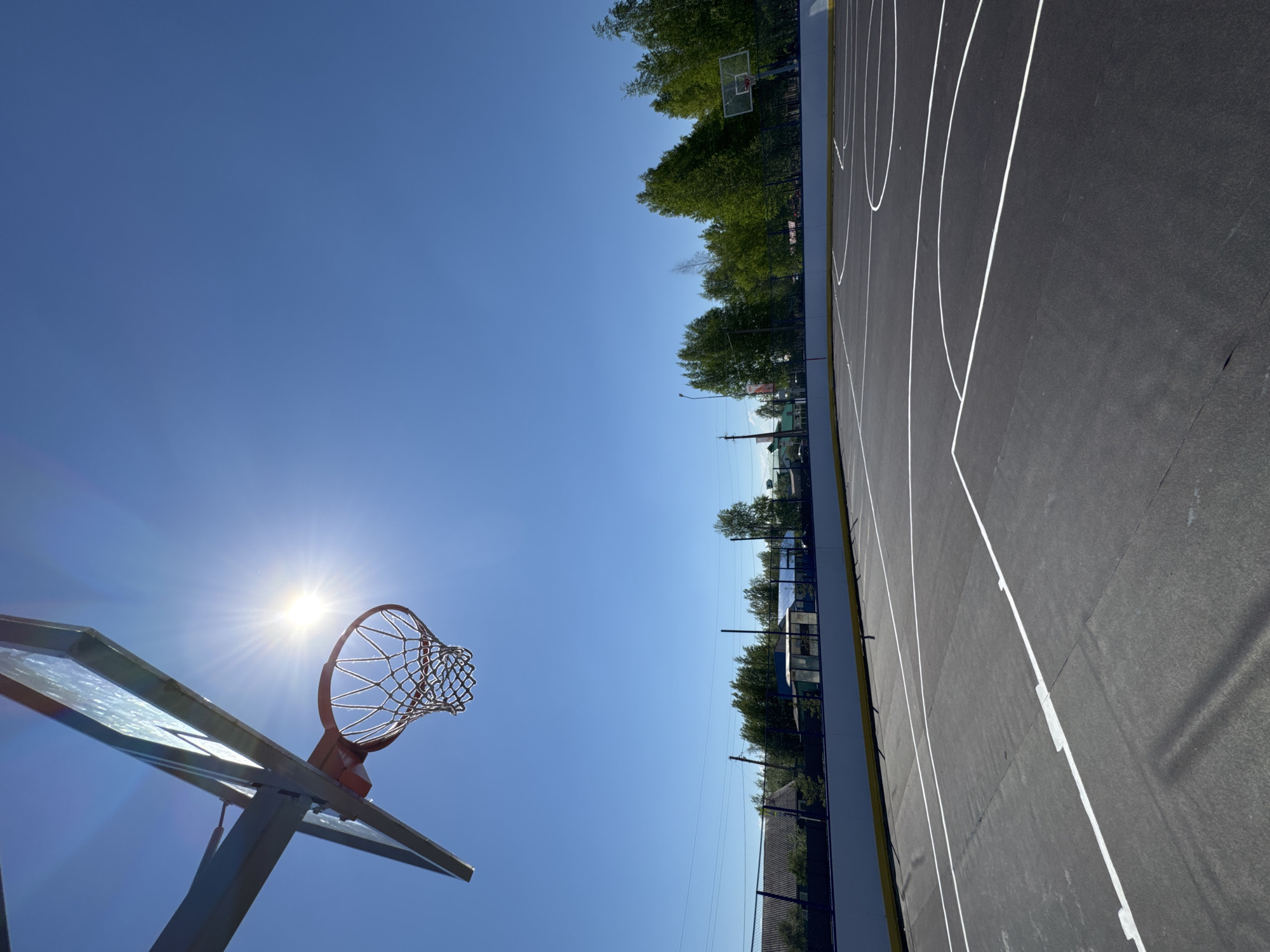 The media file presents an outdoor basketball court located in Высокий, Russia, under a clear, bright blue sky. The image is taken from a low angle, looking upwards. In the foreground, positioned to the left, is a basketball hoop featuring a transparent backboard, an orange rim, and a white net. The sun is directly behind this hoop, creating a strong lens flare and a prominent starburst effect in the sky.
The dark grey or black asphalt court, marked with crisp white lines defining the playing area, stretches from the foreground towards the background on the right side of the frame. Along the mid-ground, a line of lush green trees forms a natural backdrop behind the court, with a dark fence visible at their base. Another basketball hoop is partially discernible in the distance, near the tree line on the far side of the court. Faint structures, possibly buildings, are visible on the distant horizon beyond the trees, and a few utility poles with wires can be seen in the mid-ground on the right.
No people are present in the image, indicating the court is currently empty. The intense sunlight and cloudless sky suggest it is either midday or early afternoon. There is no visible text within the media file.