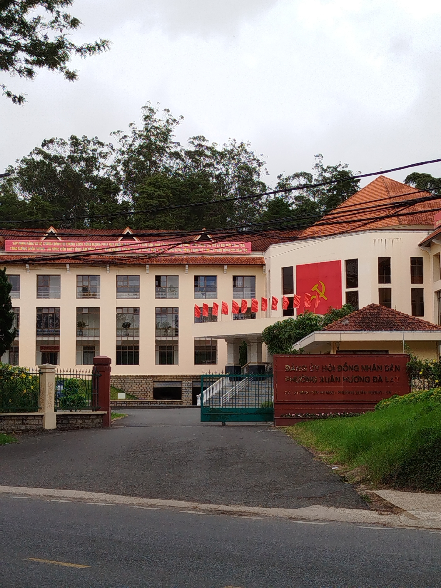 The image shows the "Đảng ủy Hội đồng Nhân dân Phường Xuân Hương Đà Lạt" building, which translates to "Party Committee of the People's Council of Xuan Huong Ward, Da Lat." The building has a large red flag with a hammer and sickle, the symbol of communism, displayed on its facade. A banner with Vietnamese text is visible above the windows of the building. The architecture appears to be a mix of colonial and Vietnamese styles, with a tiled roof and a light-colored facade. In front of the building is a sign with the name of the organization in Vietnamese. A paved road leads up to the building entrance, and trees and greenery surround the area. The sky is overcast.