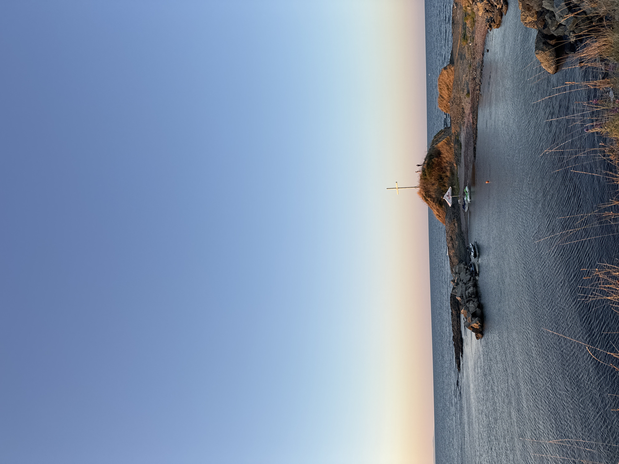 The image shows a coastal scene at sunset in Lozenets, Bulgaria. In the foreground, tall dry grasses are visible. The sea occupies the lower half of the image, with ripples on its surface. A small, rocky cove is present on the right side of the frame, with more dry grasses and rocks. In the middle ground, a small island or promontory rises from the water, topped with a large white cross and two silhouetted figures. The island also has a white umbrella and two jet skis parked on its shore. A small orange buoy floats in the water near the island. The horizon is visible in the distance, where the sea meets the sky. The sky transitions from a pale orange and pink at the horizon to a clear, pale blue at the top of the image, indicating either sunrise or sunset. The overall atmosphere is calm and serene.
