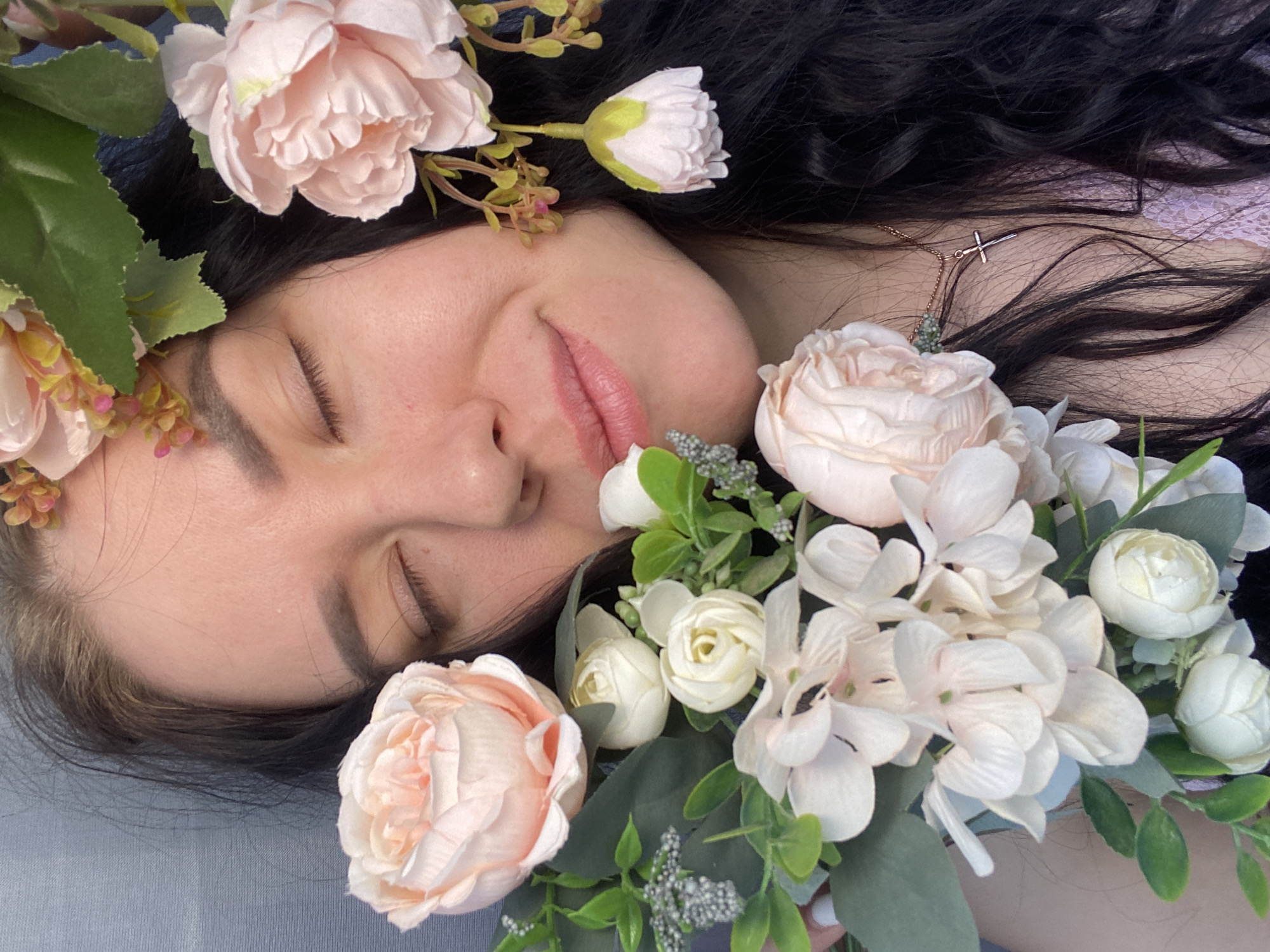 A woman with dark hair and closed eyes lies amongst peach and white flowers. She is wearing a necklace with a small cross pendant. The background is a light grey color. The location is Chaikovsky, Russia.