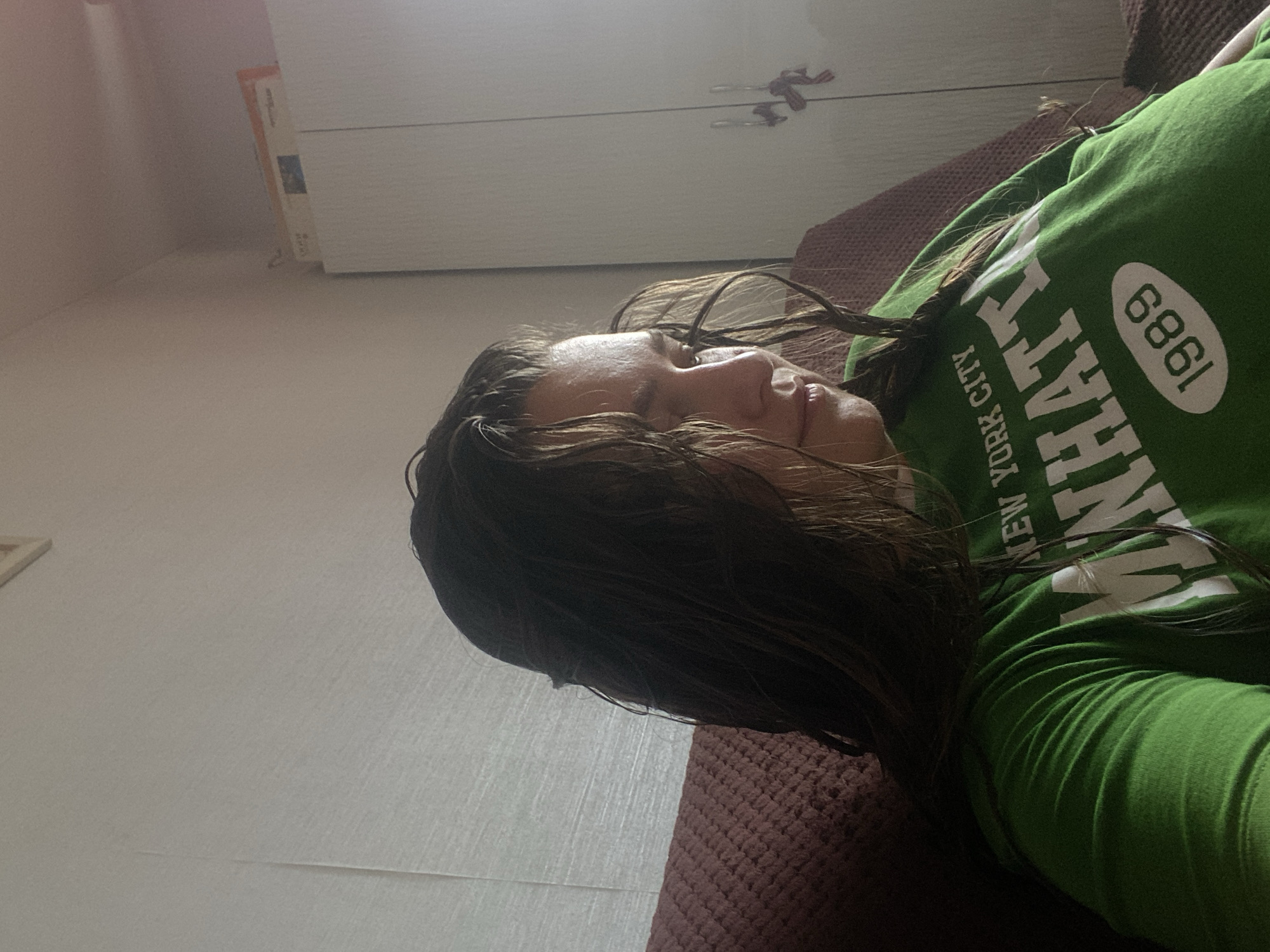 A young woman with wet, dark hair is sitting on a maroon couch. She is wearing a bright green t-shirt with white text that reads "NEW YORK CITY MANHATTAN 1989". The background appears to be an interior room with a textured, light-colored wall and a white wardrobe. There are some objects on top of the wardrobe, including what appears to be a framed picture or document. The lighting suggests it is daytime, with light coming from the left side of the frame. The location is identified as the city of Perm, Russia.