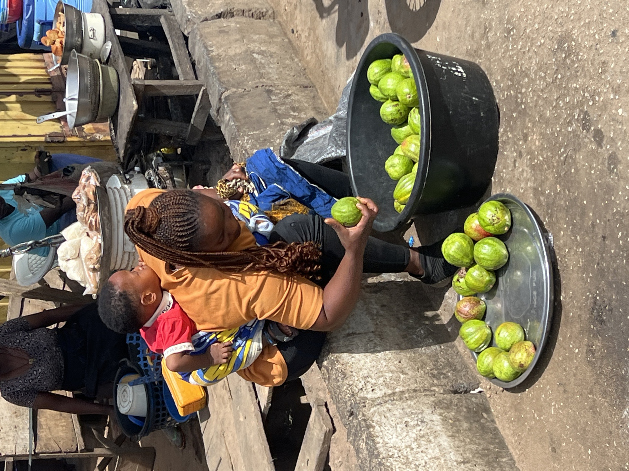 The image depicts a bustling outdoor market scene, likely in Jos, Nigeria, under bright daylight. In the foreground, a woman wearing an orange top and black trousers, with braided hair, is seated or crouched. She is carrying a baby, dressed in a red shirt and patterned shorts, in a fabric carrier on her front.
The woman appears to be a vendor, holding a round, green fruit in her right hand. Beside her, a large black plastic basin overflows with more of these green fruits, possibly guavas or a local variety. A smaller metal tray on the ground also contains several of these fruits. To the left, another woman in a dark patterned dress is partially visible. Further back, a person in a blue top is seated.
Behind the main woman, a makeshift market stall constructed from wooden planks supports various metal pots and containers, some with lids. One pot appears to hold fried dough balls or similar food items. A bicycle wheel is partially visible in the upper right. The ground is a rough concrete or paved surface, suggesting a well-trafficked area. The scene conveys a sense of everyday commerce and life, with the mother tending to her child while engaged in her work as a vendor. No discernible text is visible in the image.