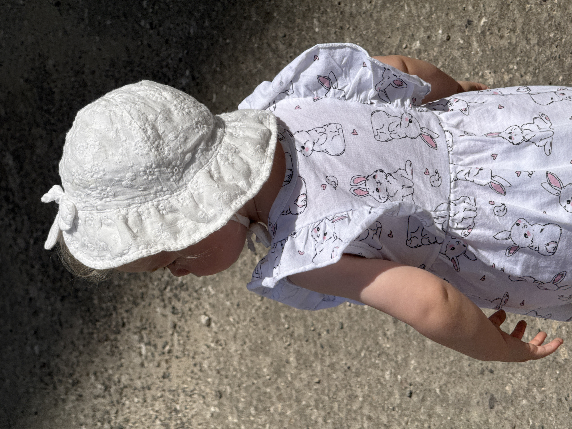 A young child, seen from the back, is wearing a white bonnet with a floral embroidered pattern and a matching white dress adorned with a print of cartoon rabbits and hearts. The child stands on a surface of pebbles and dirt, suggesting an outdoor environment. The lighting indicates it is daytime, with a shadow falling across the background. The image is a close-up shot focused on the child's attire. The specific location in Vladimir, Russia is not discernible from the visual information provided.