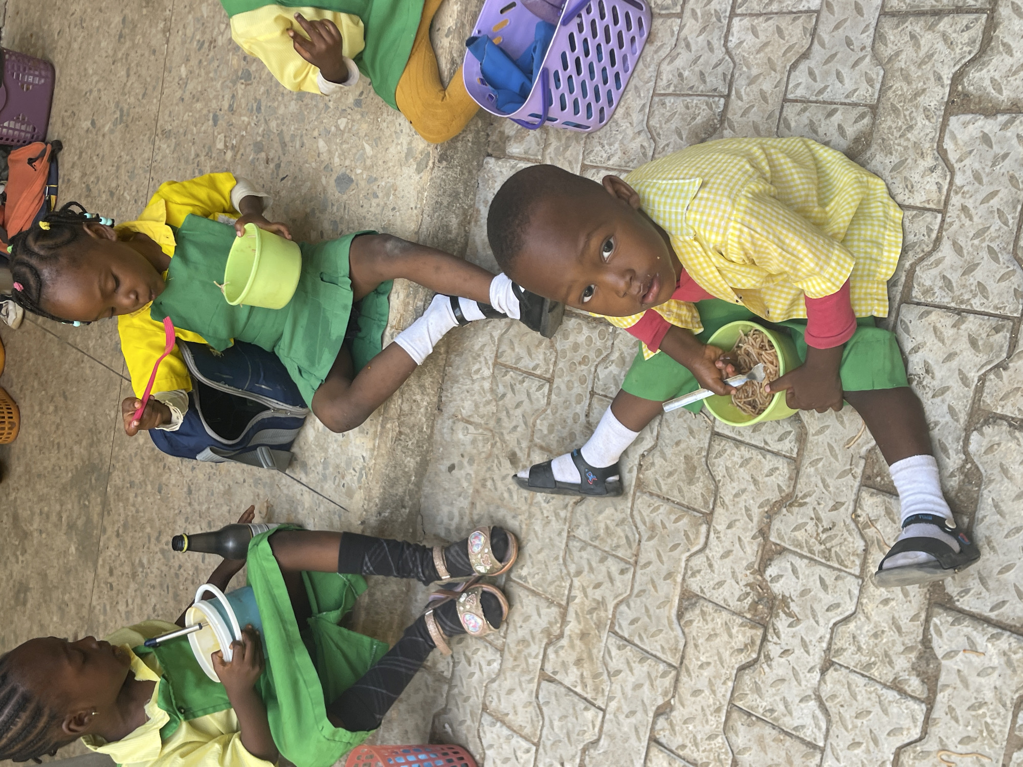 The scene depicts three children sitting on a textured concrete surface, likely an outdoor area in Akwanga, Nigeria. The children are dressed in matching school uniforms consisting of yellow shirts and green skirts or shorts. All three children are eating from light green bowls. One child, facing the camera, is actively eating with a fork. Another child appears to be resting or eating with less energy. The third child is drinking from a cup using a straw. A dark glass bottle is placed near that child's legs. A blue and purple plastic basket, a backpack, and other miscellaneous items are scattered around the children. The overall impression is one of a lunch break at school.