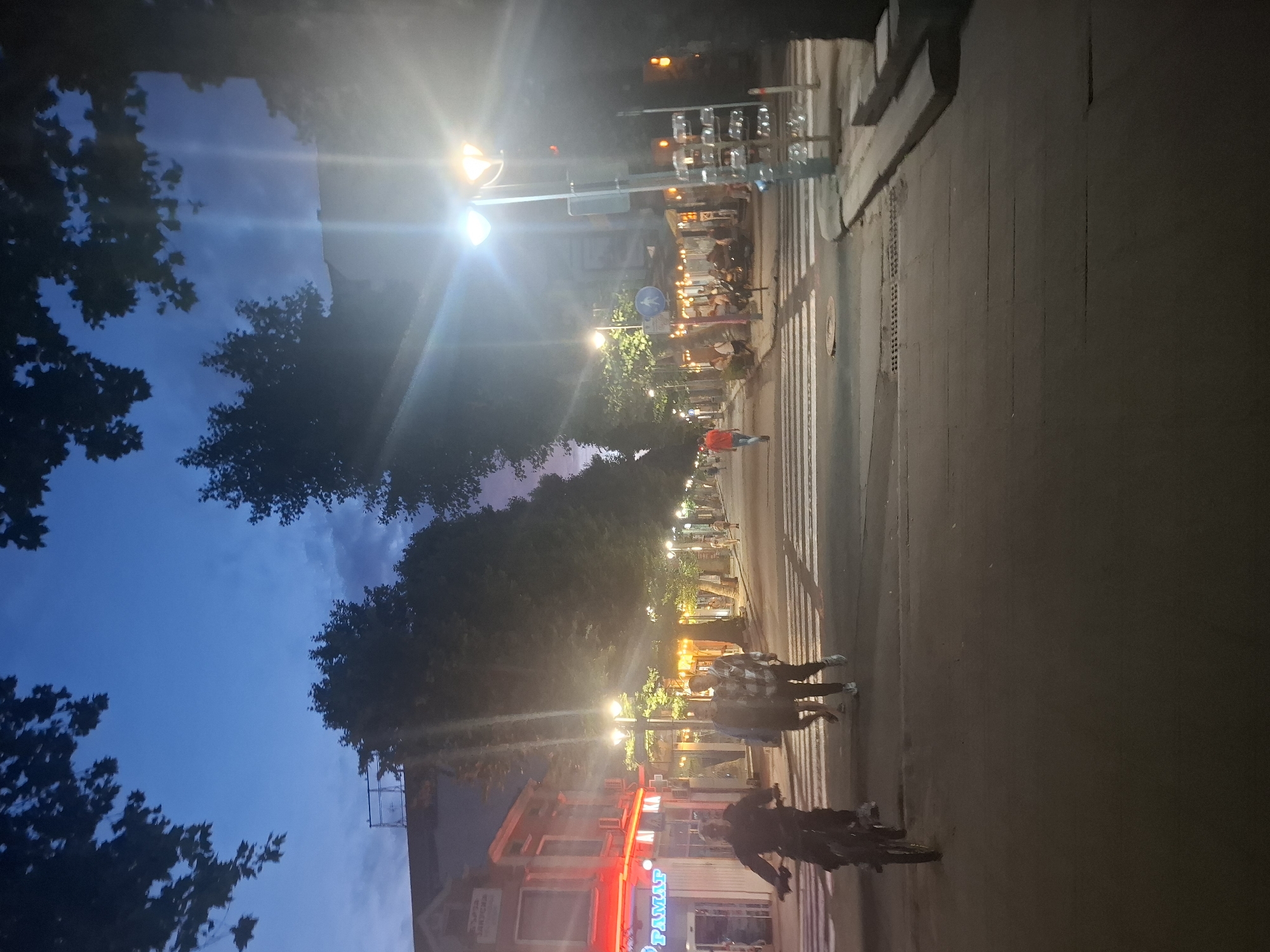 A nighttime street scene in an urban environment shows pedestrians and a cyclist on a road with a crosswalk. The sky is a deep blue, indicating dusk or early night. Streetlights and illuminated storefronts cast a warm glow on the pavement.
On the left side of the street, a person is riding a bicycle, heading towards the viewer. Two other individuals are walking across the crosswalk from left to right. Further down the street, more people can be seen walking and sitting at outdoor seating areas of establishments. A person in an orange shirt is walking away from the camera in the middle of the street.
The buildings lining the street appear to be commercial establishments, with visible signage and outdoor lighting. Trees are present along the sides of the street, some with dark foliage silhouetted against the sky. The overall atmosphere is one of a lively, illuminated street scene at twilight.