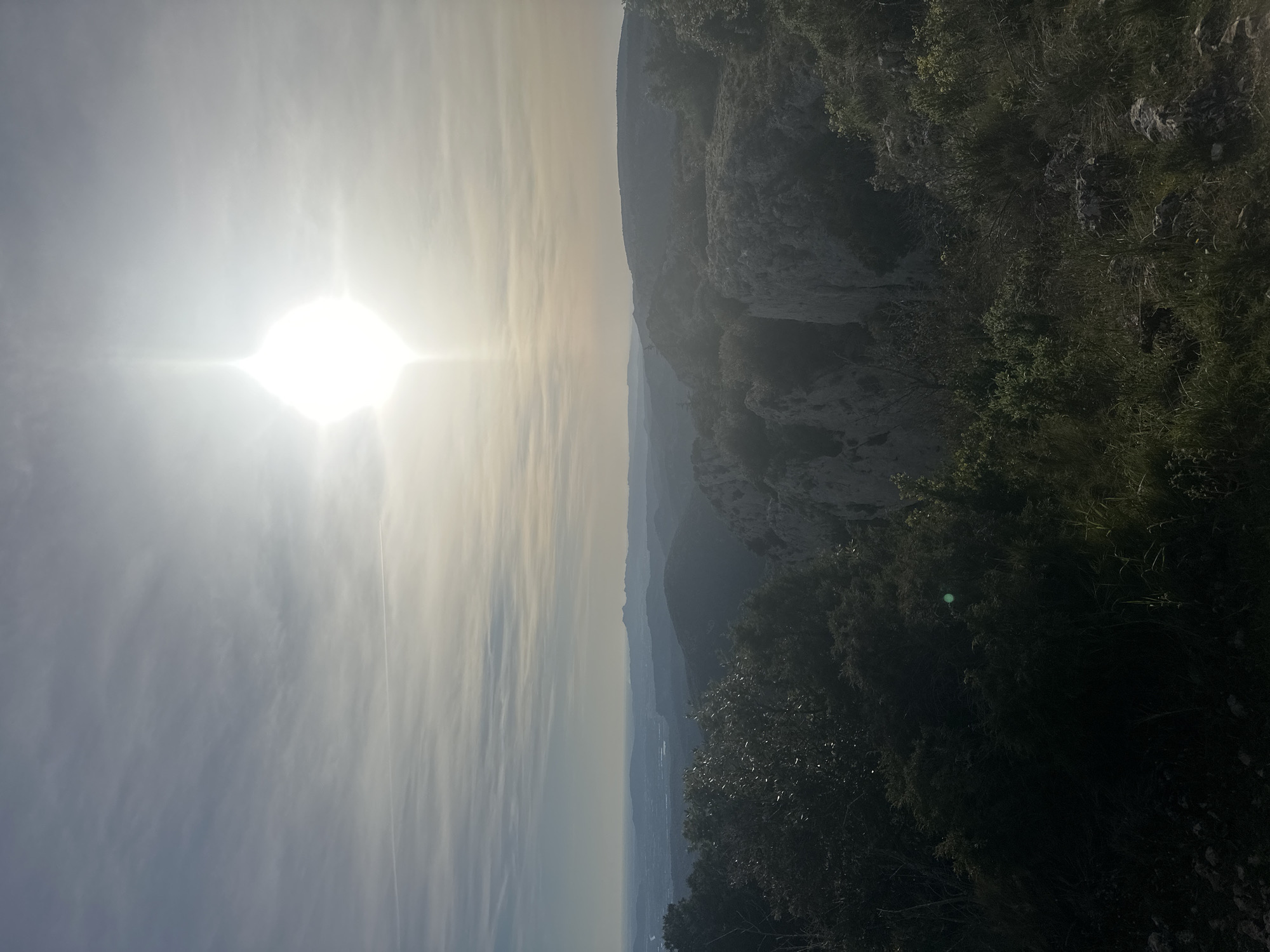 The image shows a landscape near Bonnieux, France. The sun is positioned high in the sky, dominating the left side of the frame with its bright light. Clouds are scattered thinly across the sky. To the right, a rocky cliff face descends, covered in vegetation. In the distance, there are layers of mountains fading into the hazy atmosphere. In the bottom of the image, vegetation obscures the view, which indicates that the image was taken from a high vantage point. There are no people present.