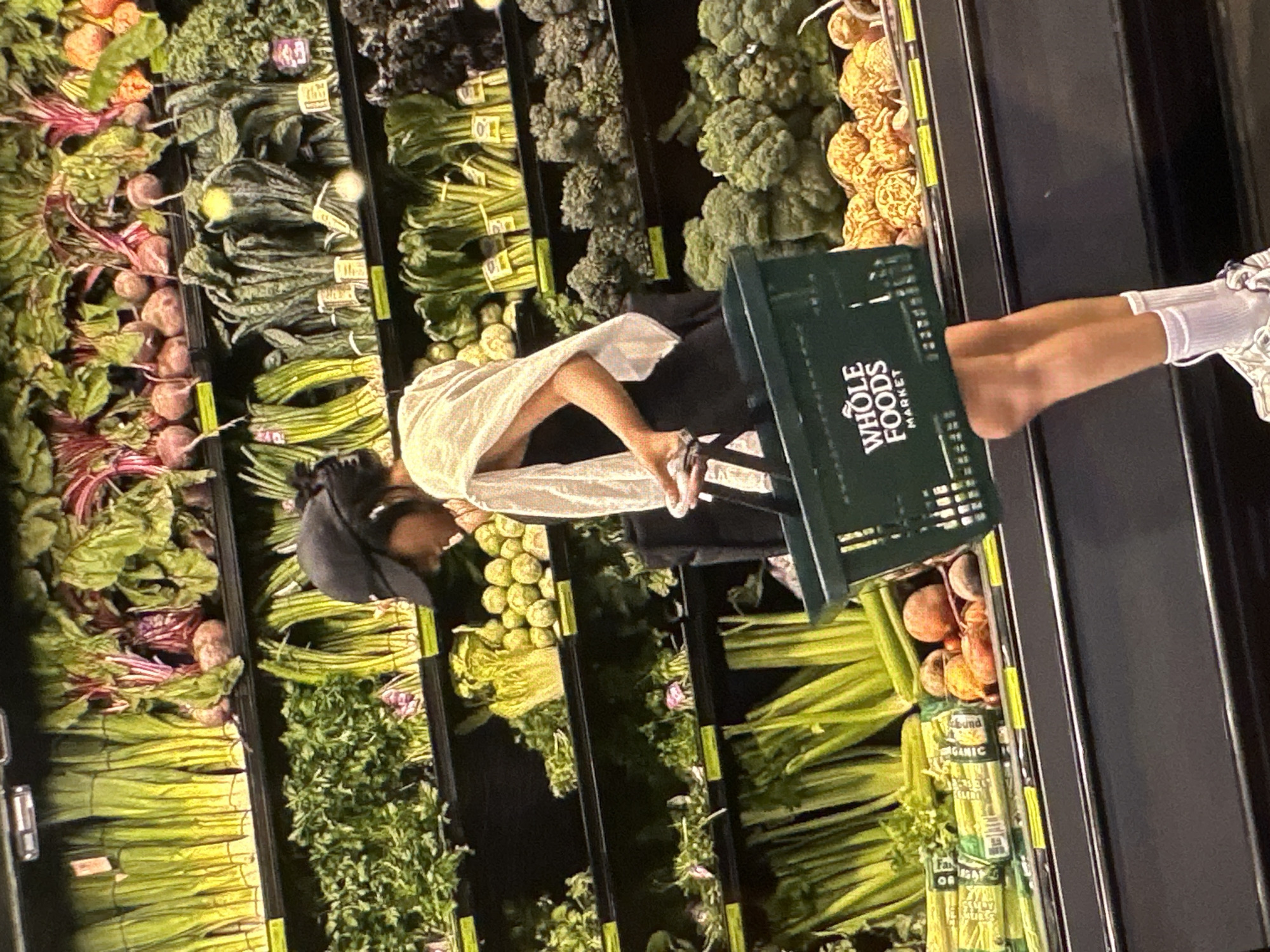 A woman wearing a black cap, white shawl, and white top is standing in a grocery store, possibly a Whole Foods Market, holding a green shopping basket with the "WHOLE FOODS MARKET" logo. The basket appears to contain fruits or vegetables. The woman is standing in front of a produce section with a variety of vegetables, including beets, kale, broccoli, celery, and cauliflower. The image is rotated and appears to be taken in a retail setting, specifically a grocery store. Yerevan, Armenia, is not visually evident in the scene.