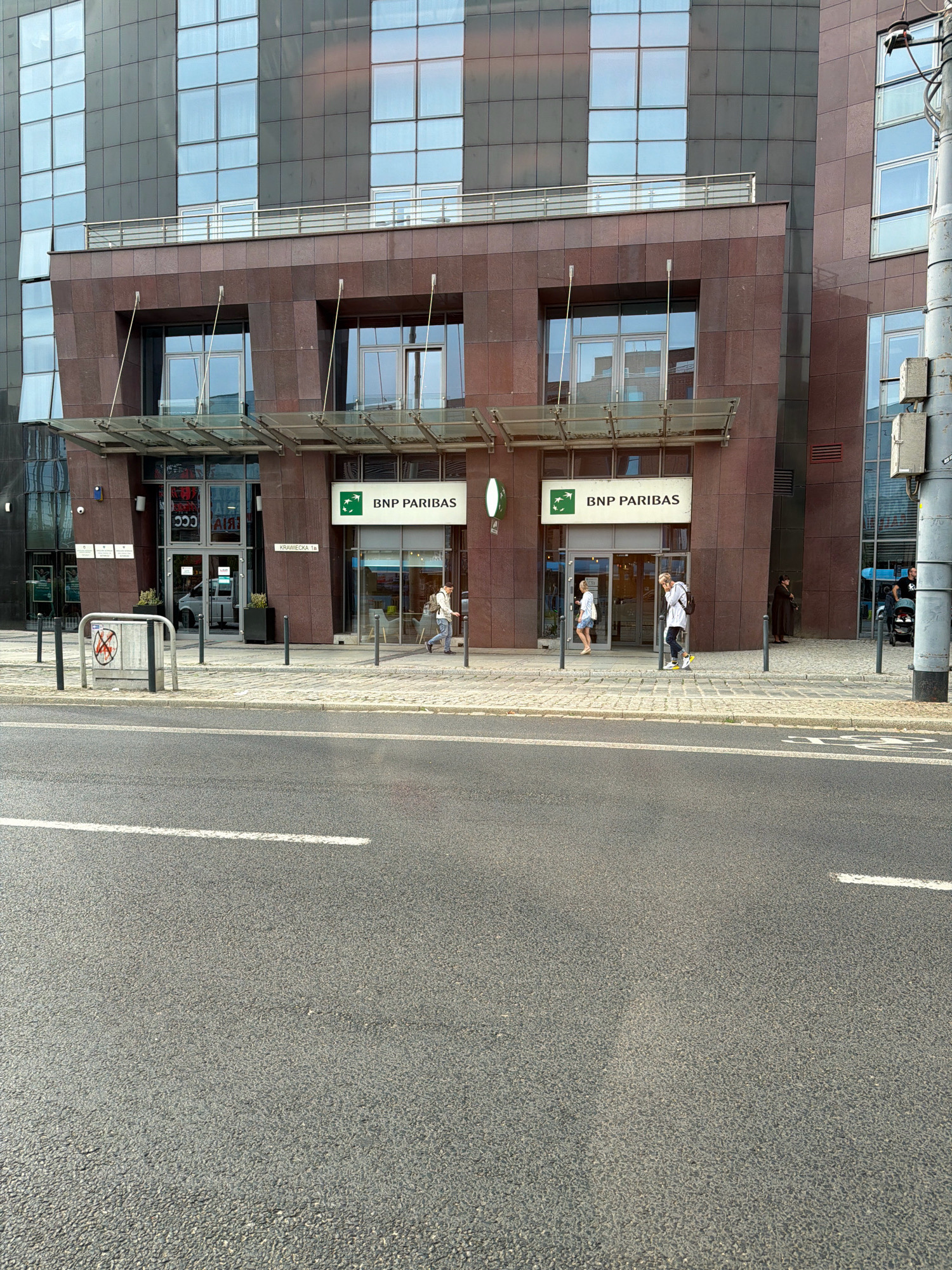 The image depicts the exterior of a modern office building, likely a bank, located in Wrocław, Poland. The building features a facade with dark, polished stone and large, reflective windows. In front of the building, a paved sidewalk runs along a street with asphalt road markings.
Several individuals are visible on the sidewalk. On the left, two people are near the entrance of the building, one of whom is entering. To the right, a person wearing a white top, dark pants, and a backpack is walking away from the building, touching their face. Further to the right, another individual in dark clothing stands near a stroller, and a person in dark attire is partially visible near the entrance.
Key elements include:
* **Building:** A contemporary multi-story building with a dark, stone-clad lower section and large glass windows above. The lower section has a prominent canopy over the entrance.
* **Signage:** A green and white sign displays "BNP PARIBAS" with the bank's logo. Another sign indicates "KRAWIECKA 1b," suggesting the street address.
* **Street and Sidewalk:** A paved sidewalk separates the street from the building. The street is asphalt with visible white lane markings.
* **Objects:** A metal bike rack or barrier with a circular sign containing a red cross is positioned on the sidewalk. Planters with green plants are located near the entrances. A utility pole with attached boxes stands on the right side of the frame.
* **People:** Multiple individuals are present, engaged in various activities such as walking and entering the building.
The scene appears to be a typical urban environment on a bright day, with no overt signs of distress or unusual activity. The presence of a bank and office building suggests a commercial district.