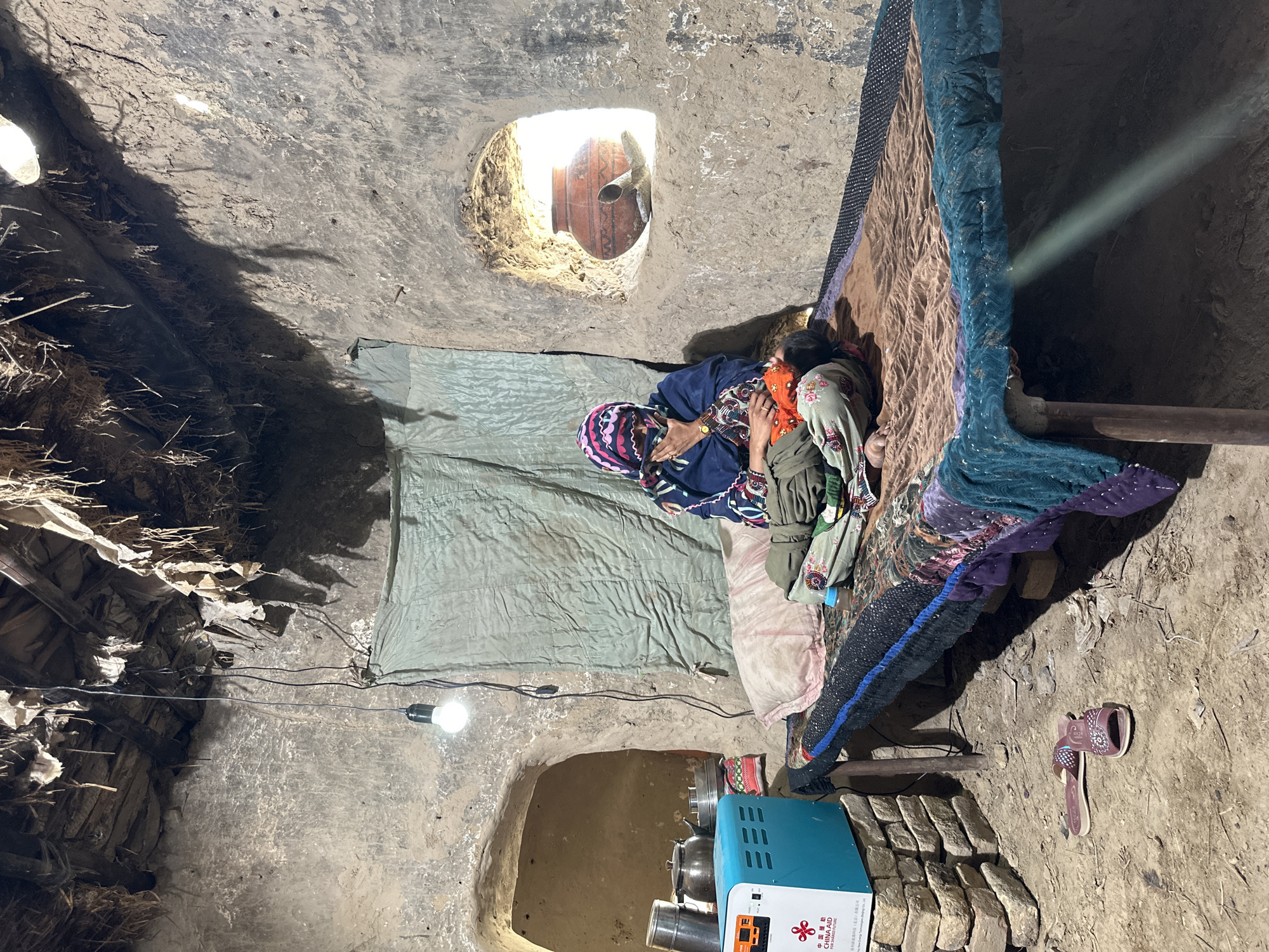 A woman rests on a bed in an earthen-walled room. She is adorned in colorful traditional clothing and headwear. The room is sparsely furnished, with a light bulb hanging from the ceiling. The room has a dirt floor and the walls appear to be made of packed earth. A generator is positioned in the lower right corner of the room. Light enters the room from two small, circular openings in the ceiling. The scene is located in the city of Quetta City Tehsil, Pakistan.