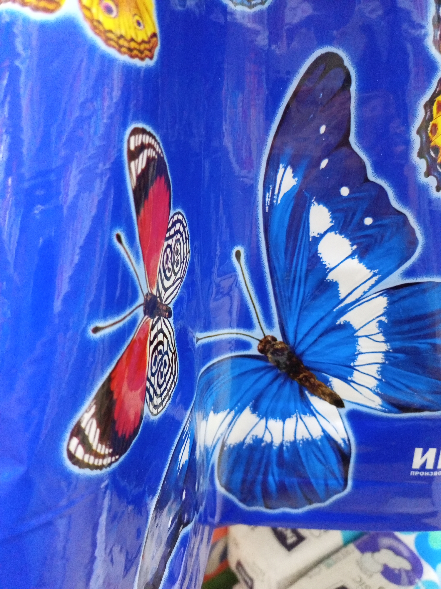 The image displays a close-up of a bright blue plastic bag adorned with colorful butterfly decals. Two prominent butterflies are visible: a red and black butterfly with intricate white patterns on its wings is positioned to the left, and a large blue butterfly with white markings is to the right, appearing larger and closer to the viewer. The texture of the plastic bag is evident, with some creases and reflections visible. In the lower right corner, a partial view of text in Russian, "И ПРОИЗВО", is present. Below the bag, the blurry forms of stacked white and blue packaged goods suggest this might be a retail environment or storage area. The image does not provide any information about the city of Kevsala, Russia, nor does it contain any individuals or specific actions.