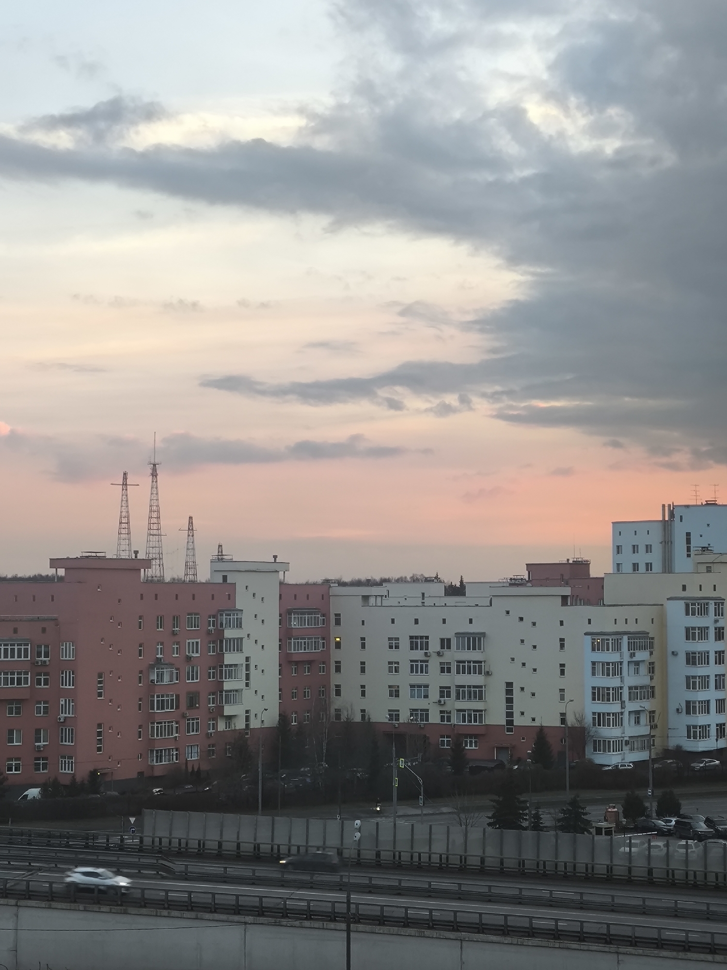 The image captures a cityscape at dusk in Khimki, Russia. Several multi-story apartment buildings of varying colors (pink, white, yellow, and blue) dominate the middle ground, with numerous windows and some balconies visible. Behind the buildings, three lattice-like radio or electrical towers stand out against the colorful sky. The sky transitions from light pink near the horizon to gray clouds higher up. In the foreground, a highway runs horizontally, with blurred cars suggesting movement. A gray concrete barrier separates the highway from the viewer's perspective. Further back, a road runs parallel to the highway, with cars and trees interspersed.