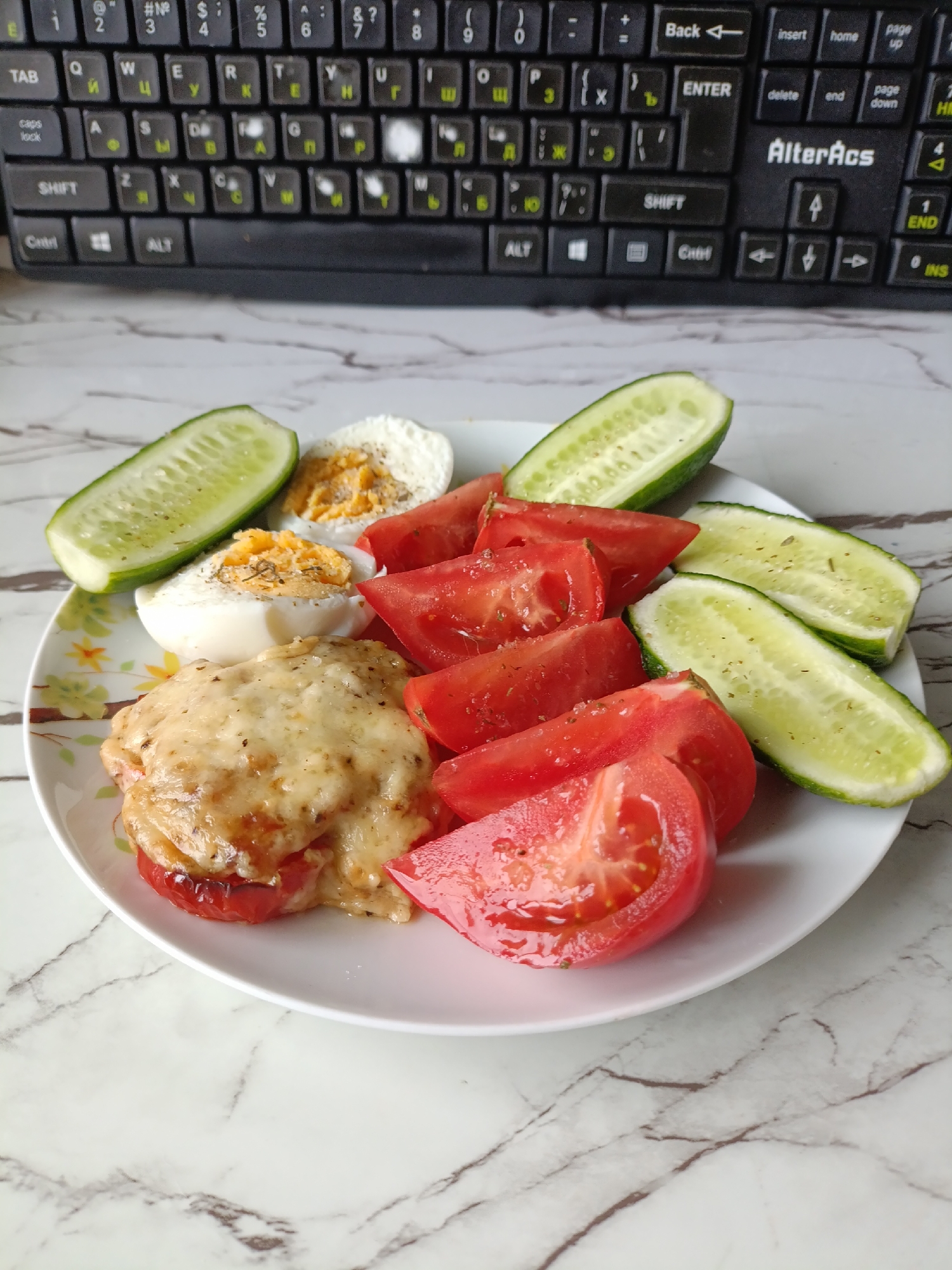 A close-up, overhead shot shows a white plate with food on a white marble surface, with a portion of a black keyboard in the background. The plate contains a piece of chicken topped with melted cheese, sliced tomatoes, two halves of a hard-boiled egg, and slices of cucumber. The food is seasoned with herbs and spices. The keyboard appears to be a standard QWERTY layout with Cyrillic characters visible. The scene is well-lit, with the focus on the food.
