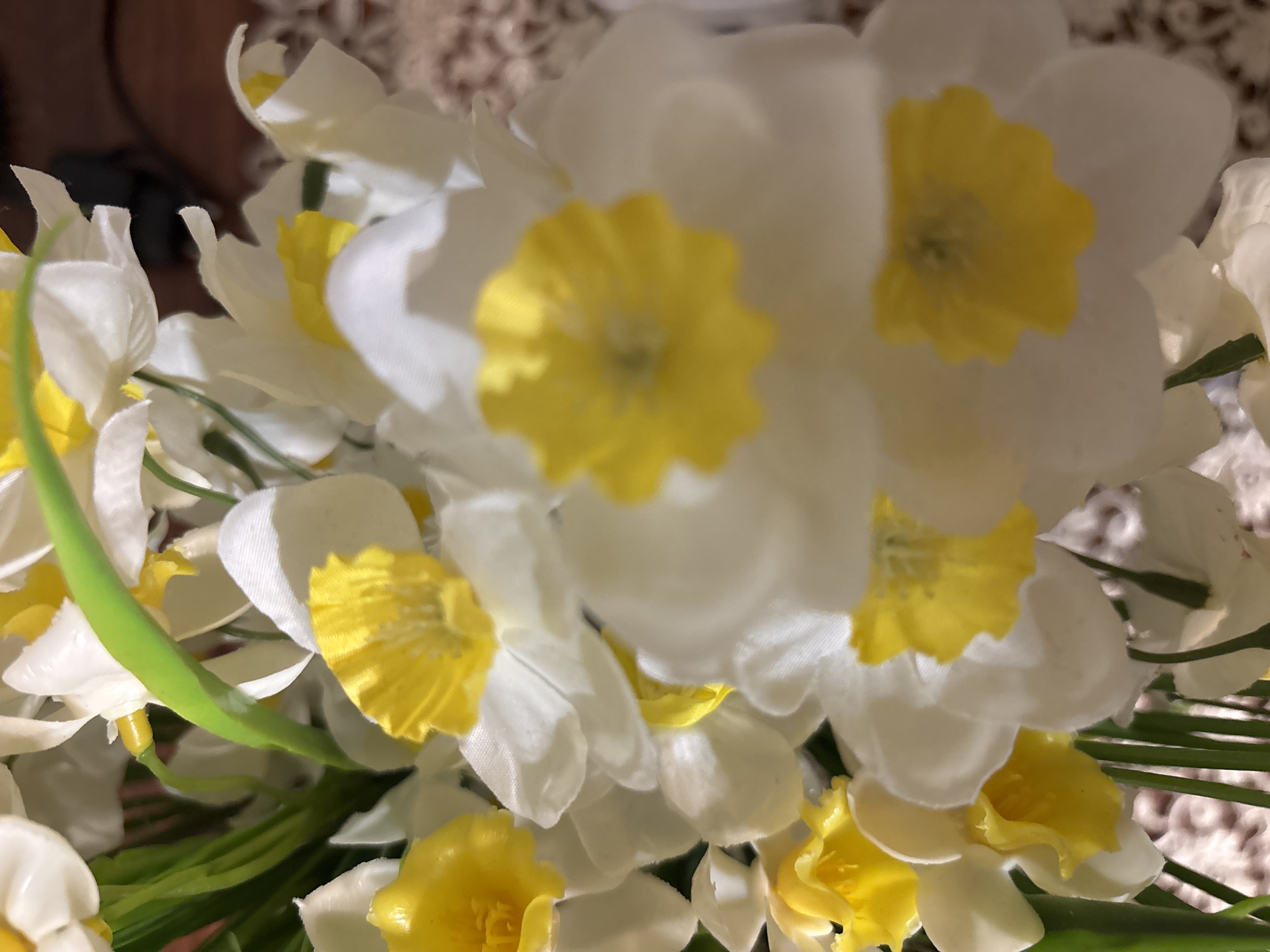 This image captures a close-up view of a vibrant bouquet of artificial flowers, likely daffodils or narcissus. The dominant colors are white and bright yellow, with accents of green.
The flowers are densely arranged, filling the majority of the frame. Each flower features crisp white petals surrounding a distinct, ruffled yellow corona (the central cup-like structure). Several of these yellow centers are prominent, some appearing slightly blurred due to the shallow depth of field, while others offer a clearer view of their intricate textures. The white petals show subtle folds and creases, characteristic of fabric artificial flowers.
Green stems are interspersed among the blossoms, adding a natural contrast to the white and yellow. The lighting appears soft and diffused, highlighting the delicate textures of the petals and giving the overall arrangement a fresh and cheerful appearance.
The background is softly out of focus, suggesting an indoor setting and emphasizing the flowers as the primary subject. No people or discernible activities are visible. There is no visible text within the image. The image is a static shot of a floral arrangement.