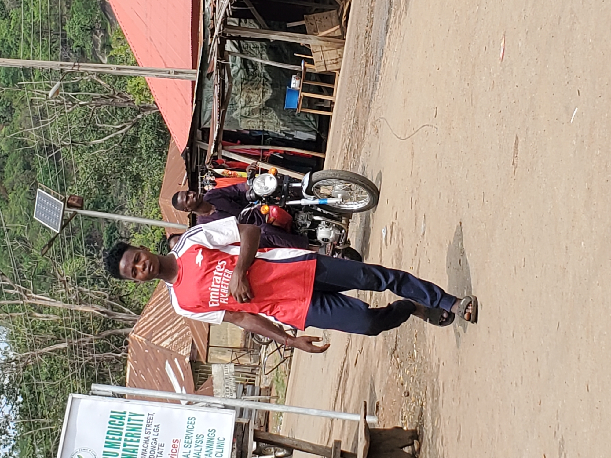 The image shows a man in a red and white Arsenal Emirates FLY BETTER jersey, standing on a dusty road in Mararaba, Nigeria. He is wearing dark pants and sandals. Behind him is a motorcycle with a rider. A sign for IBU Medical Maternity is visible to the left, indicating it offers medical services, analysis, family planning, and clinics. The environment appears to be a street scene with buildings and trees in the background. Overhead power lines are also visible.