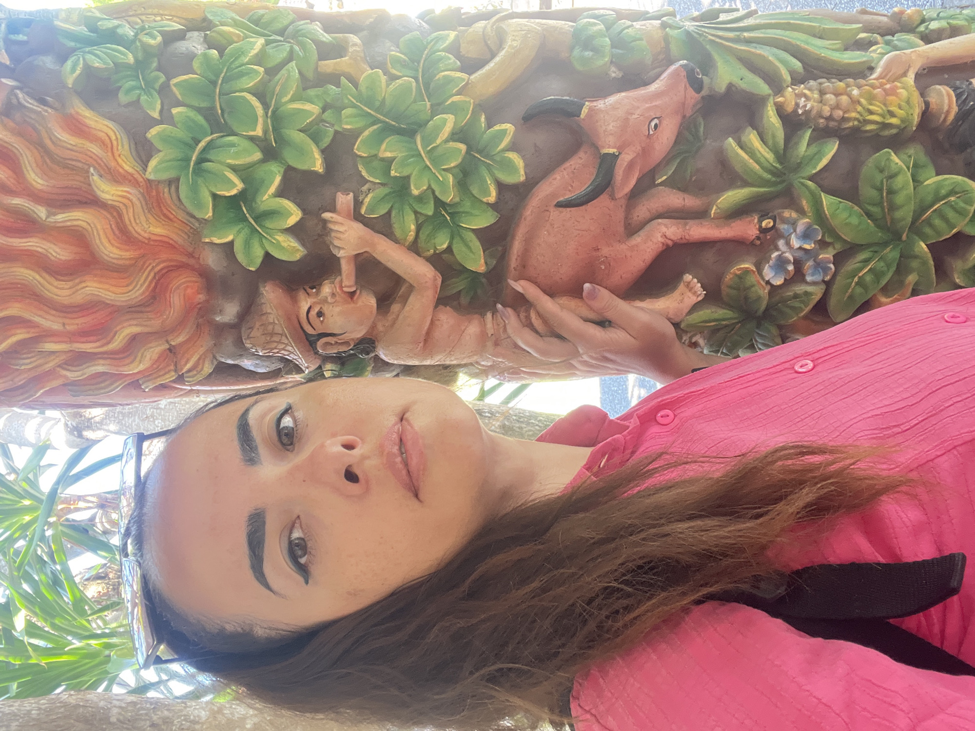 In Kuta Selatan, Indonesia, a young woman with dark hair and a pink collared shirt stands next to a carved pillar. She is looking at the camera, her sunglasses perched on her head. The pillar features a carving of a person wearing a hat and playing a flute, with a bull beneath them. The carving also depicts flames, leaves, and corn. The environment is outdoors, with palm trees visible in the background.