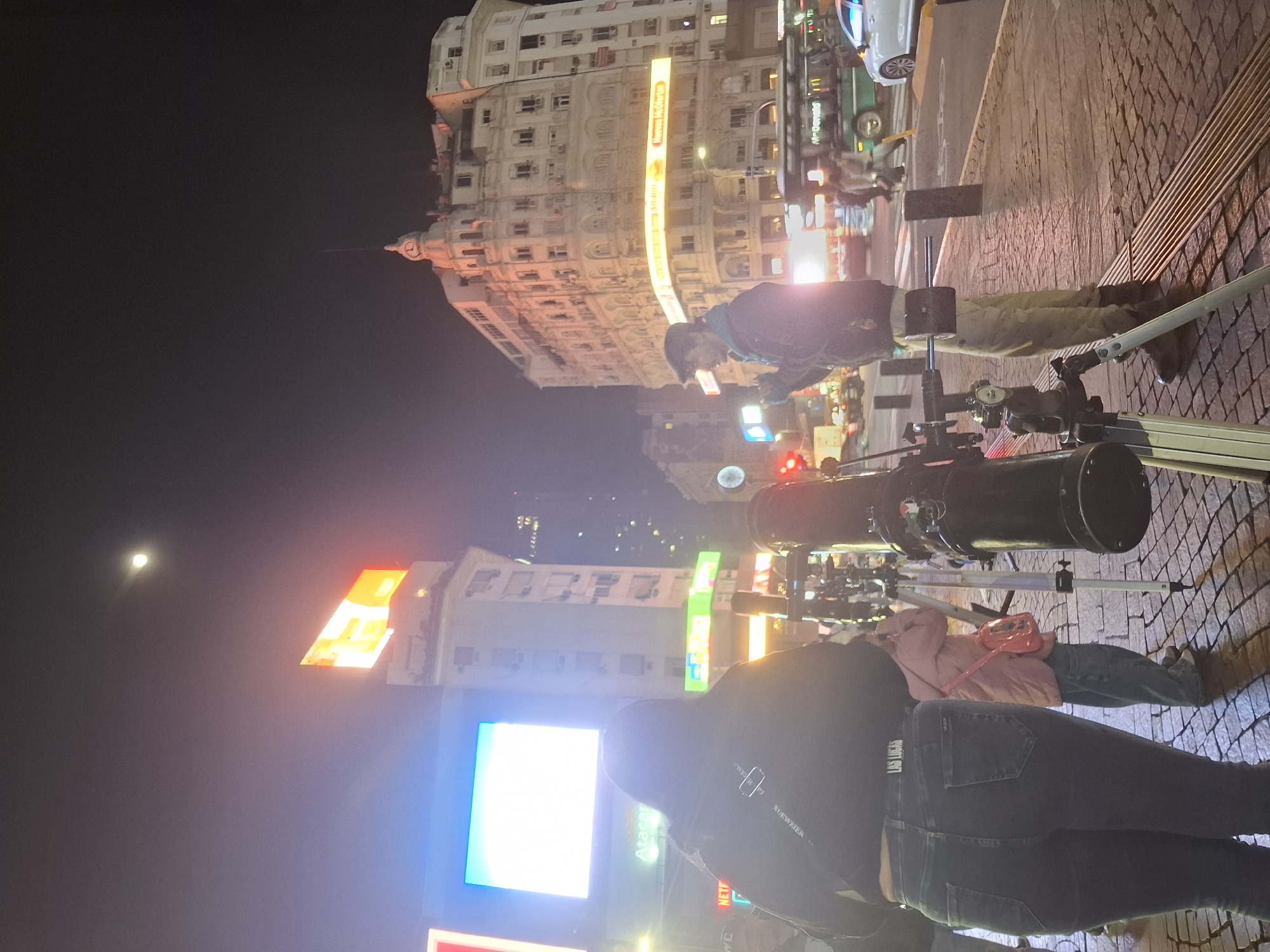 A street scene at night shows several people gathered around telescopes. A tall, ornate building stands in the background on the left, with illuminated signs. A full moon is visible in the upper-left of the image. The scene appears to be in a city, judging by the presence of buildings, streetlights, and pedestrians. The image shows a telescope on the street pointing toward the moon. The location is the city of Buenos Aires, Argentina.
