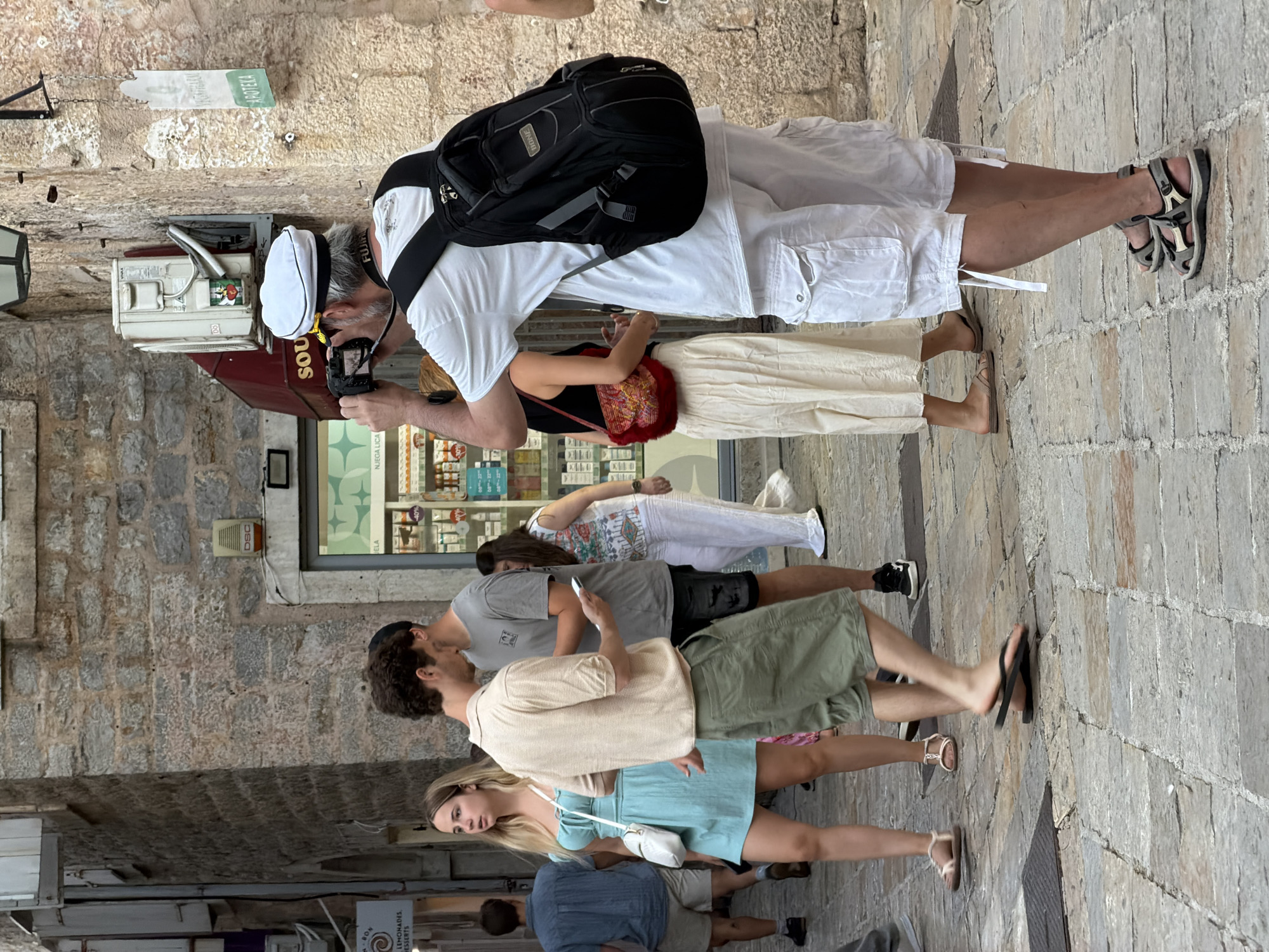 The image depicts a street scene in Budva, Montenegro. A man wearing a white cap, t-shirt, and cargo shorts is holding a camera to his face, appearing to take a photograph. He has a black backpack on. In front of him, a woman in a light blue dress and a man in a beige shirt and green shorts are walking. Behind them, other individuals are visible in the background, including a person in a blue shirt and shorts, a woman in a white and floral print top and white pants, and another woman in a black top and cream-colored skirt with a red purse. The environment consists of stone buildings with narrow windows and doorways, and the ground is paved with cobblestones. A shop front with shelves stocked with goods is partially visible on the left. A sign reading "APOTEKA" is mounted on the wall to the right. The overall scene suggests a busy tourist area on a bright day.