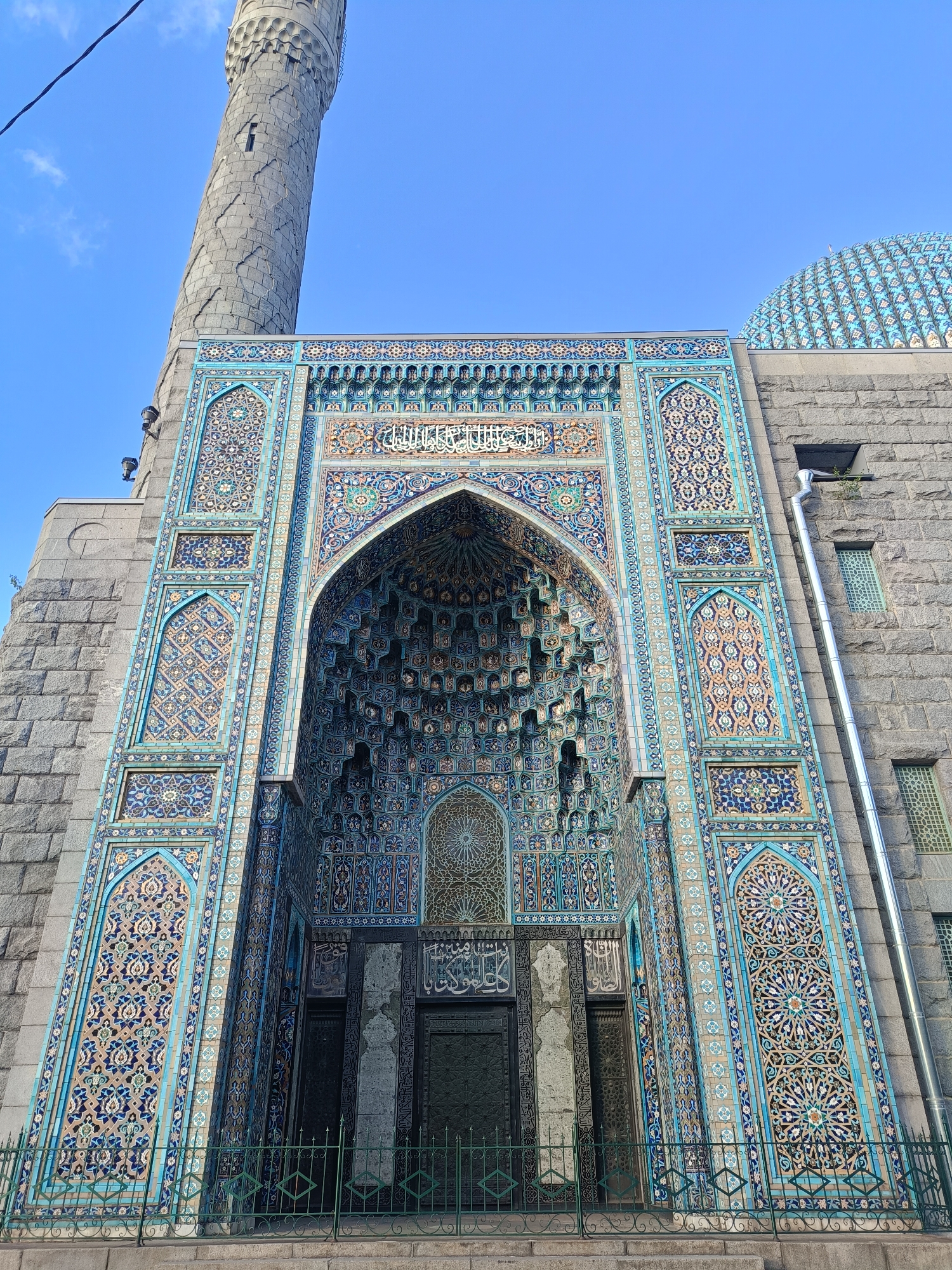 This is a low-angle, full-shot photograph of the Saint Petersburg Mosque, located in Saint Petersburg, Russia. The mosque's main entrance is the focal point, featuring an elaborate, multi-layered archway decorated with intricate blue, green, and beige tiles. The tiles display geometric patterns and Arabic calligraphy. Flanking the entrance are tall, narrow sections of similarly tiled walls. Above the entrance, a larger arched section is visible, also adorned with detailed tilework. To the left of the entrance, a portion of the mosque's tall, grey stone minaret is visible, with its distinctive textured surface. To the right of the entrance, the grey stone facade of the mosque continues, with a portion of a blue-tiled dome visible at the top right of the frame. A metal downspout runs vertically down the stone wall on the right. In the foreground, a green metal fence runs along the base of the structure, with stone steps leading up to the entrance area. The sky above is clear and blue. There are no people visible in the image. The image is captured in daylight.