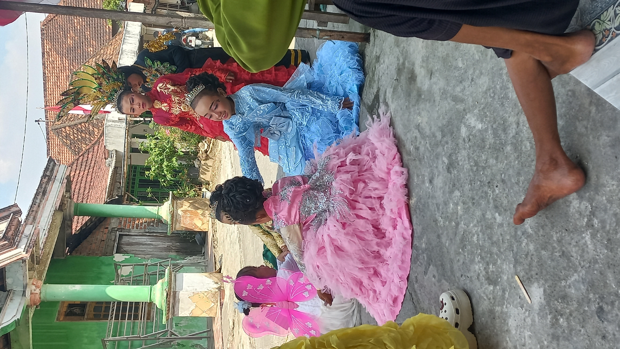 In Kedungmegarih, Indonesia, on a sunny day, a group of young girls dressed in elaborate, princess-like gowns are gathered outdoors, possibly for a performance or celebration. A girl in a bright blue dress with voluminous ruffles and a tiara smiles as she reaches out to touch the hair of another girl. This second girl is wearing a pink, fluffy, floor-length gown adorned with silver sparkles and has a tiara in her intricately styled hair. To her left, a girl in a white dress with pink butterfly wings sits, facing away from the camera. Further back, other children are visible in colorful costumes, including one in a red dress and another in a dark outfit with an elaborate, feathered headdress. The background reveals a simple, weathered building with a green facade and a tiled roof, typical of a rural Indonesian setting. The ground is paved with concrete. A pair of white platform sandals and a small wooden stick are on the ground near the seated children. In the foreground, the legs and feet of two individuals are visible, one person seated and another with their legs extended.