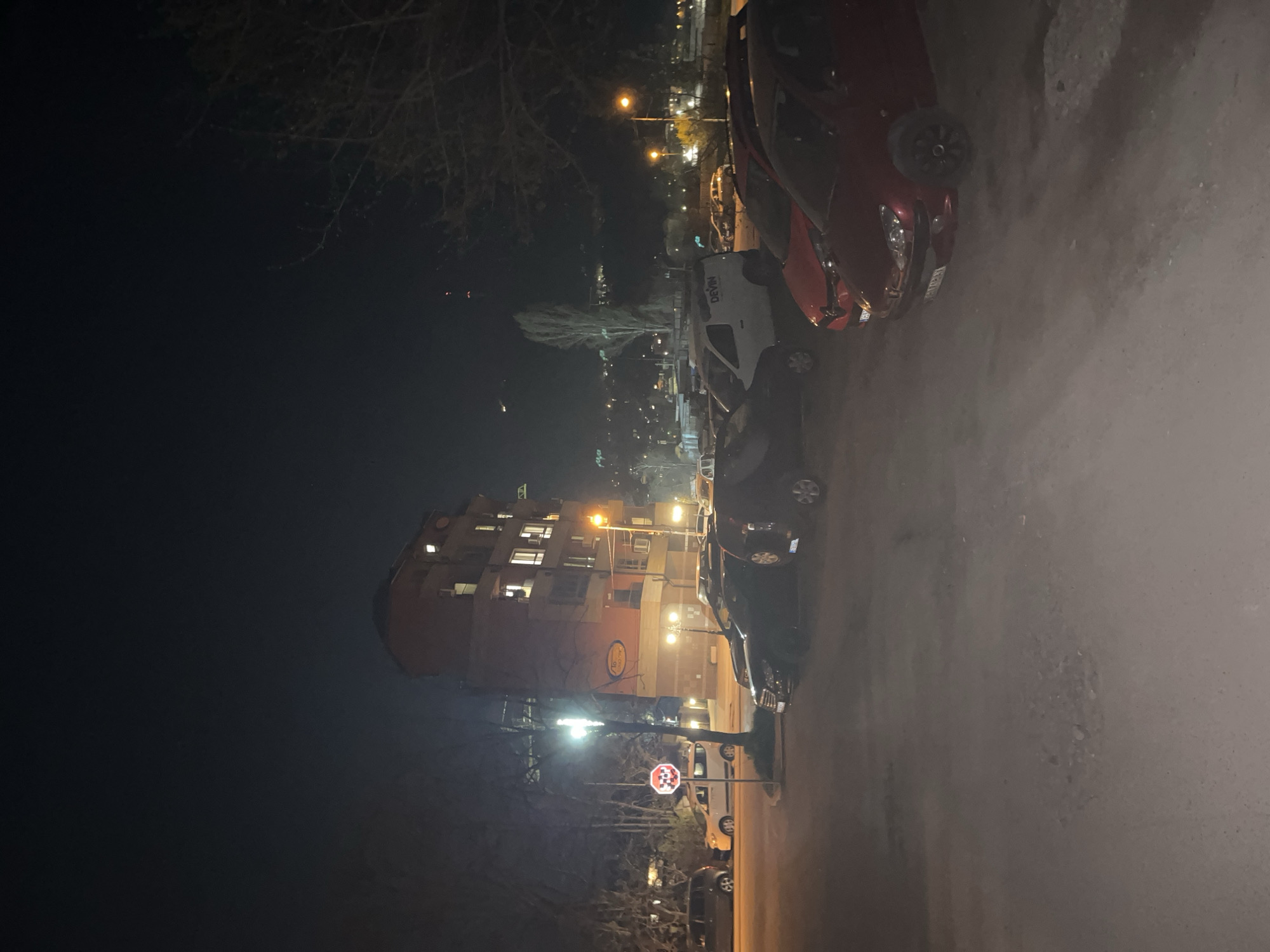 The image shows an urban street scene at night in Sofia, Bulgaria. The perspective is ground-level, with the camera angled upward towards a multi-story building, likely residential. The building appears to have multiple lighted windows, and an establishment named "Sofia Residence" is present, illuminated by external lighting. The street is lined with parked cars on the right. A STOP sign is visible near the building. The sky is dark and there are bare trees visible. Streetlights provide illumination.