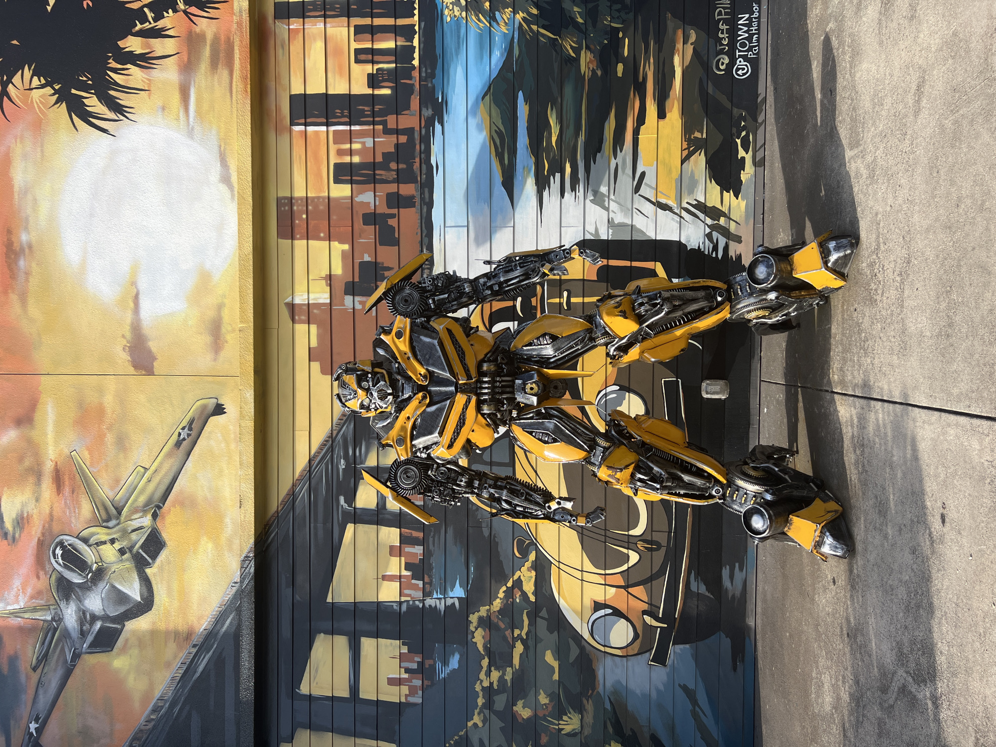 A full-body, three-quarter-angle shot shows a detailed, life-sized Bumblebee Transformer statue standing in front of a vibrant mural. The mural depicts a multi-layered urban landscape with a bridge and a fighter jet at the top, a city skyline and a yellow classic car in the middle, and a tropical sunset with palm trees on the right. The statue is predominantly yellow and black, with intricate mechanical details and exposed robotic parts. The ground in front of the statue is concrete, with visible shadows cast by the statue and the sunlight. The bottom right of the mural features graffiti tags that read "@Jeff Pihl" and "UPTOWN Palm Harbor." The image appears to be taken outdoors during the day, with bright natural lighting.
