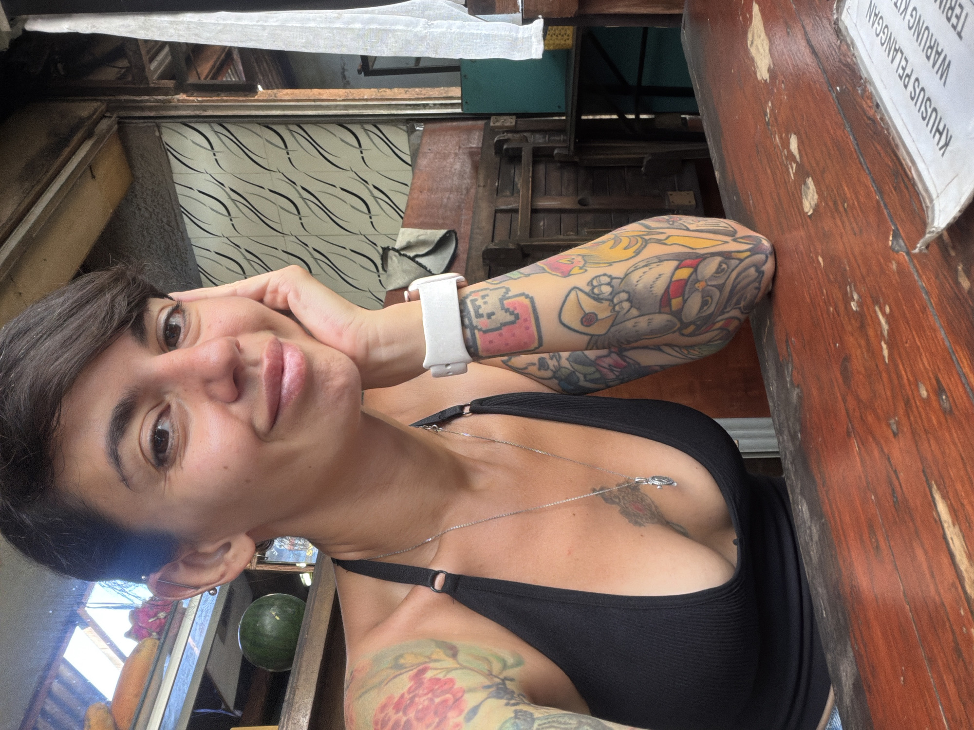 A woman with short, dark hair and tattoos on her arms is seated at a wooden table. She is wearing a black tank top and a silver necklace. Her right hand is resting on her cheek. The background shows a window with some fruit visible, and a wall with a patterned design. A white sign with text in a foreign language is on the table in the foreground. The scene appears to be an indoor/outdoor eating area.