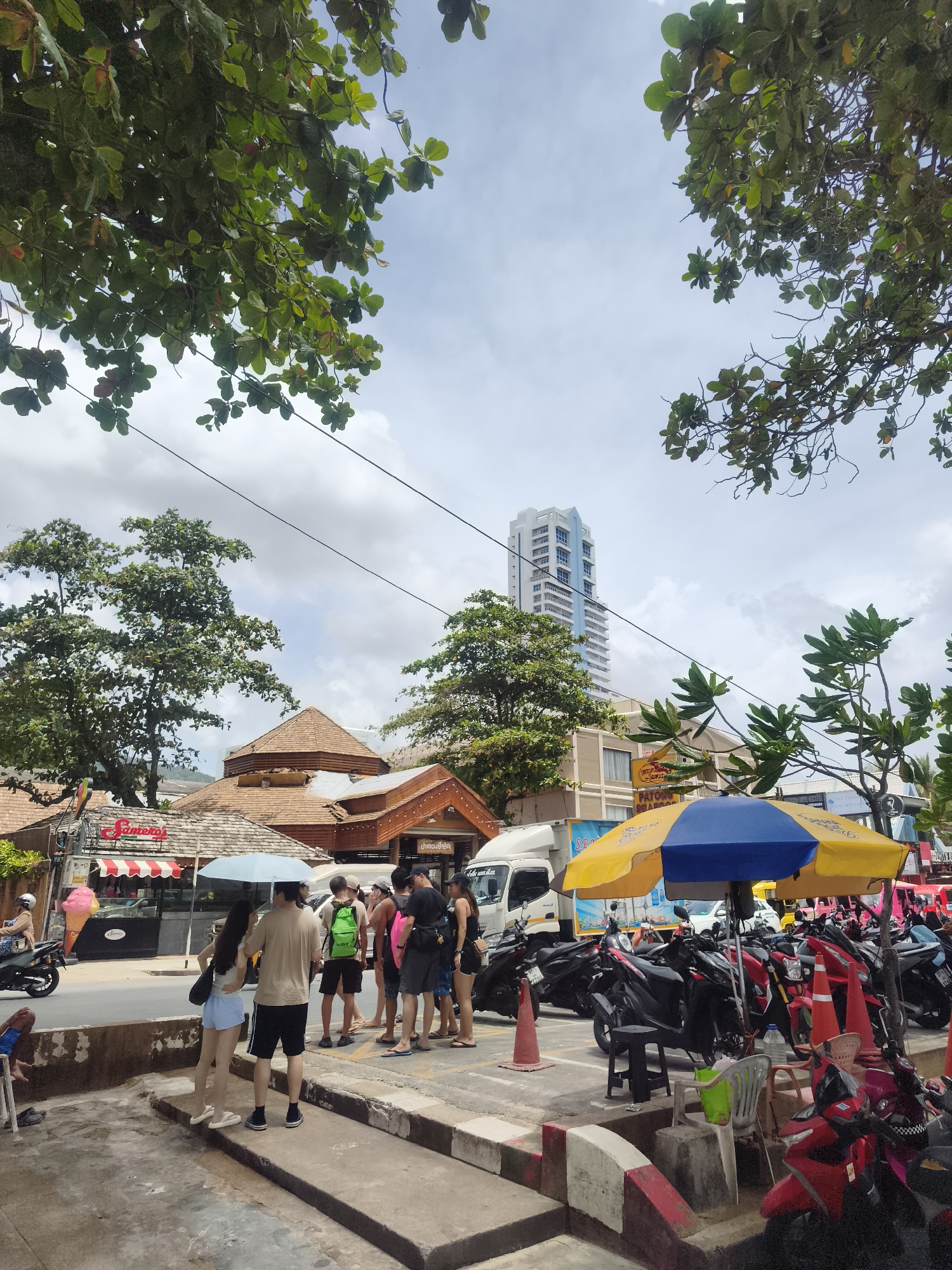 A street scene in Patong, Thailand, features a line of parked motorcycles under a yellow and blue umbrella, with a few individuals standing nearby. In the background, a tall, modern building stands out against a cloudy sky, with a few trees and smaller buildings dotting the landscape. A white truck is parked to the left of the motorcycles, and to the right, more motorcycles are visible. A couple is standing under a light blue umbrella in the foreground, facing away from the camera.