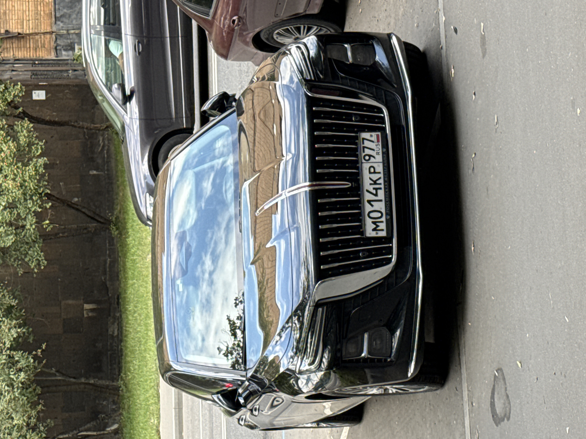 The image shows a black sedan parked on a street in Moscow, Russia. The car's license plate reads "MO 14KP 977". In the background, another dark grey sedan is visible, as well as a red-brown car. Trees and buildings line the street, and the sky is reflected in the car's windshield. The scene appears to be a sunny day.