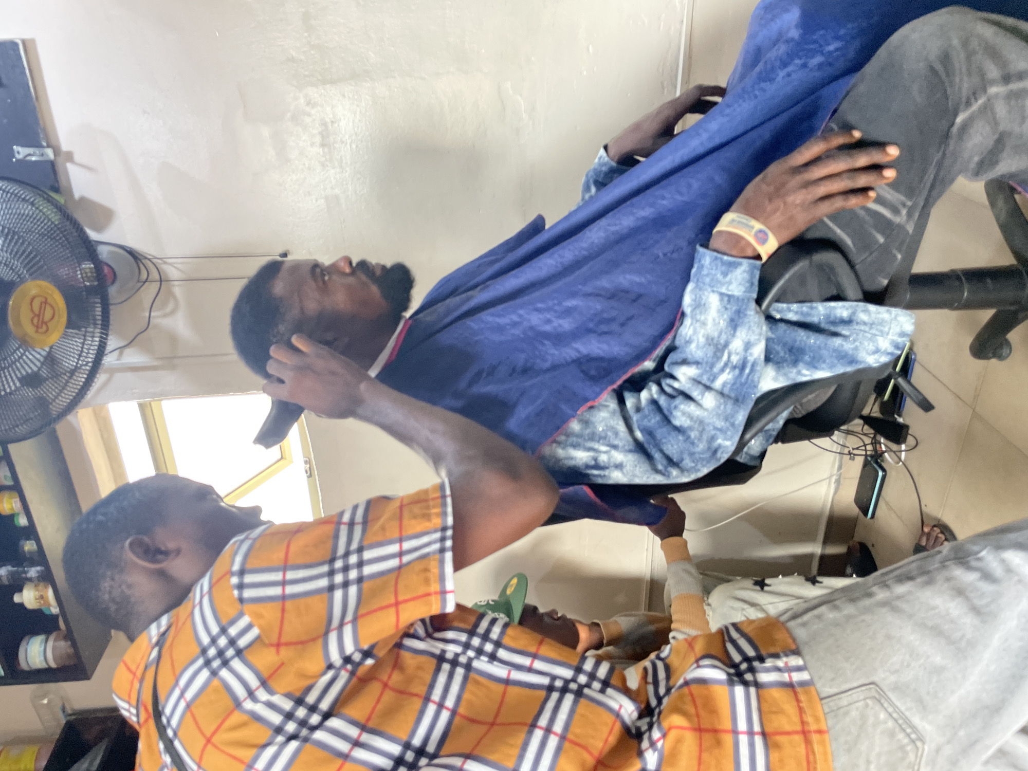In an indoor setting in Ado, Nigeria, a barber wearing an orange and white plaid shirt is giving a haircut to a man draped in a blue barber cape. The client is seated in a barber chair. A shelf containing bottled products is visible on the left side of the frame, next to a fan mounted on the wall. A window is visible in the background.