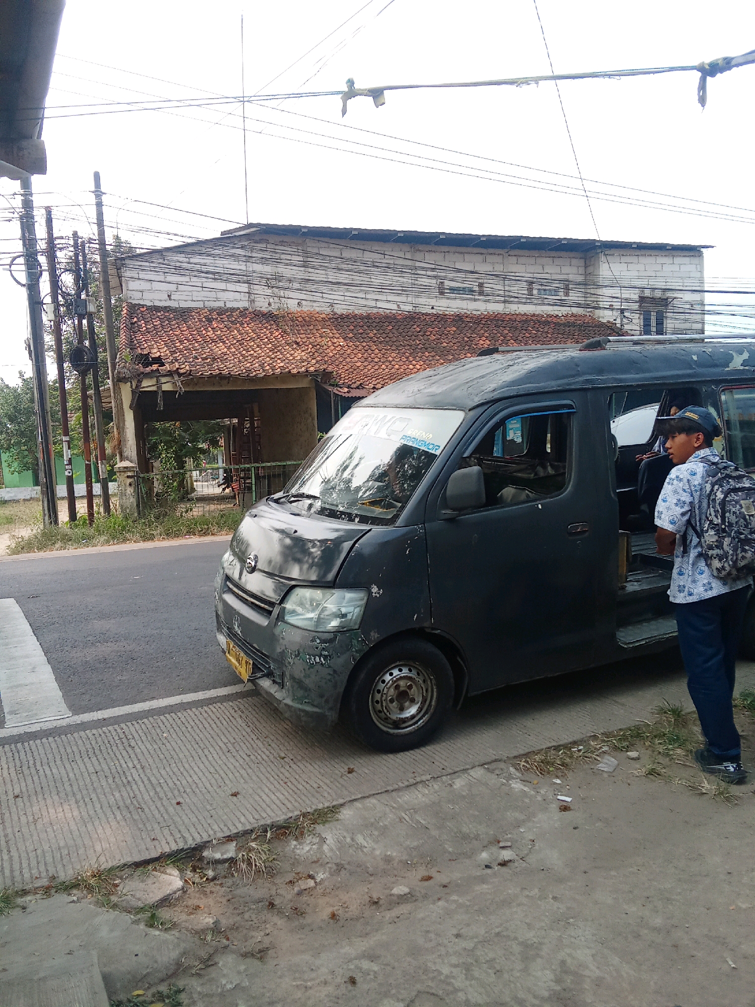 In Warunggunung, Indonesia, a dark-colored minivan is parked on the side of a street, with its passenger door open. A young male wearing a blue cap, a patterned short-sleeved shirt, and dark pants stands next to the open door, facing away from the camera, and appears to be looking into the minivan. He carries a camouflage backpack. The minivan's front is visible, showing signs of wear and tear. The street appears to be paved with asphalt, and a concrete pedestrian walkway runs alongside it. In the background, a damaged building with a tiled roof is visible, along with numerous overhead electrical wires. The environment suggests an urban or semi-urban setting.