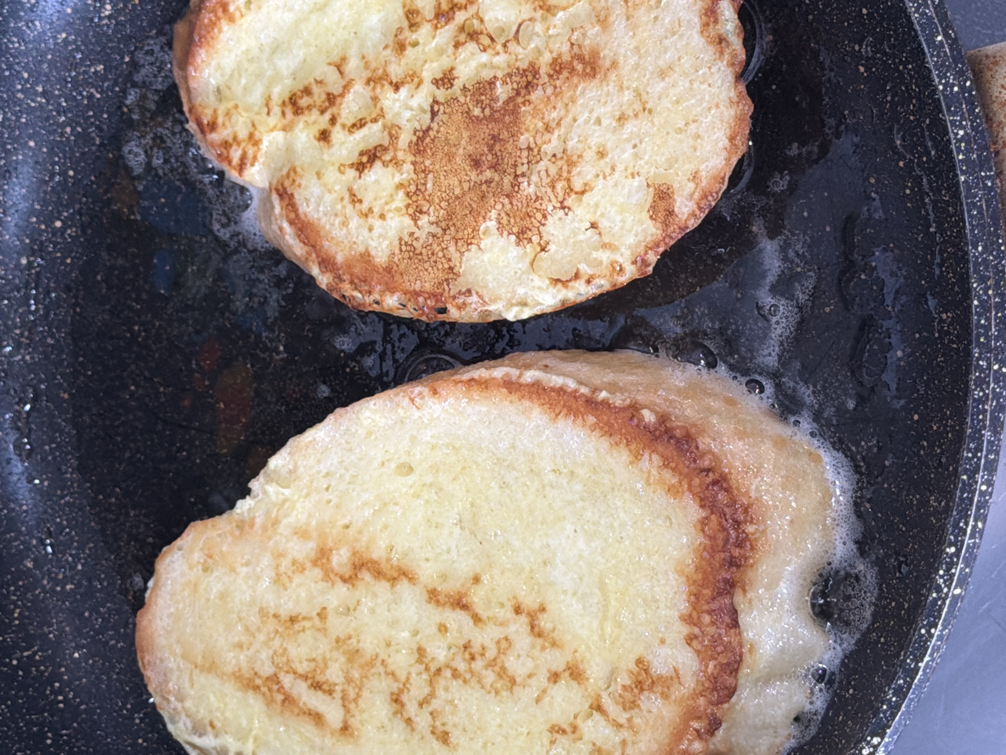 Two pieces of golden-brown French toast are cooking in a dark, speckled frying pan. The pan contains bubbling oil or melted butter, indicating active frying. The upper piece of French toast shows a more pronounced browning pattern, with shades of light yellow and darker toasted areas. The lower piece also exhibits browning along its edges and a lighter, irregular pattern across its surface. Both pieces of bread appear to be thick-cut. The scene captures a close-up, overhead view of the cooking process, focusing solely on the pan and its contents. There are no people visible, and no text is present in the image. The setting suggests an indoor kitchen, with the food being prepared.