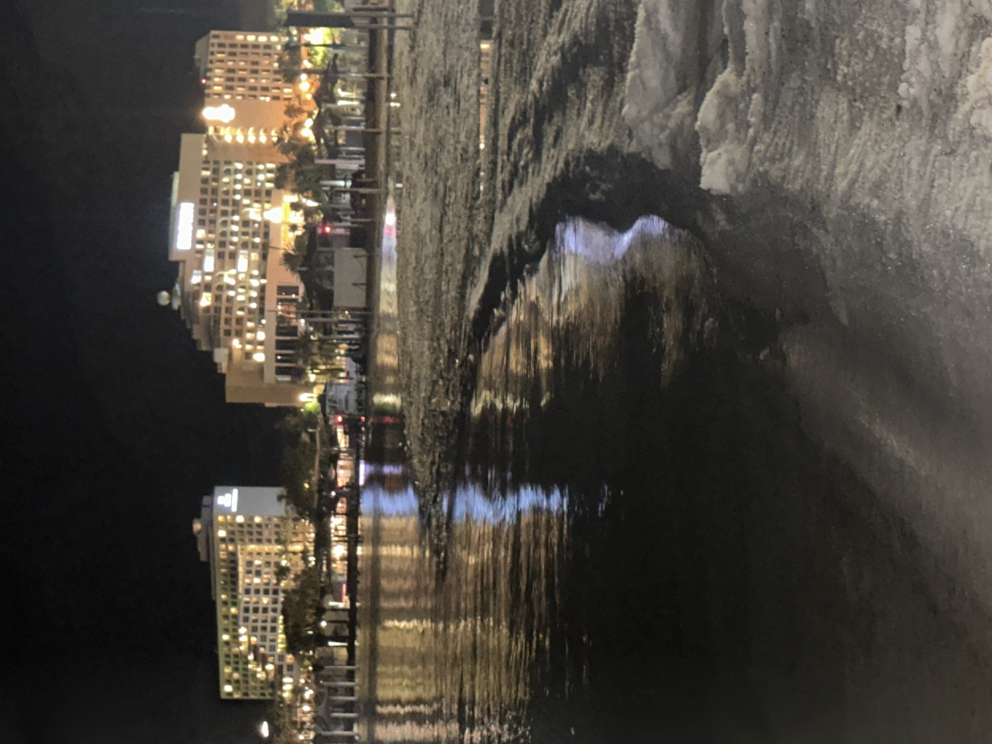 The image captures a nighttime scene on a beach in Eilat, a city in the Tamar Regional Council, Israel. The foreground features a body of water reflecting the lights from the city. The water is calm, with gentle ripples disturbing its surface. Beyond the water, a sandy beach is visible, leading to a boardwalk with several small structures and palm trees. In the background, two large hotel buildings are brightly illuminated, with many windows lit up. The sky is dark and clear, typical of a night scene. There are no people or animals visible in the image. The overall impression is one of a serene and well-lit coastal resort at night.