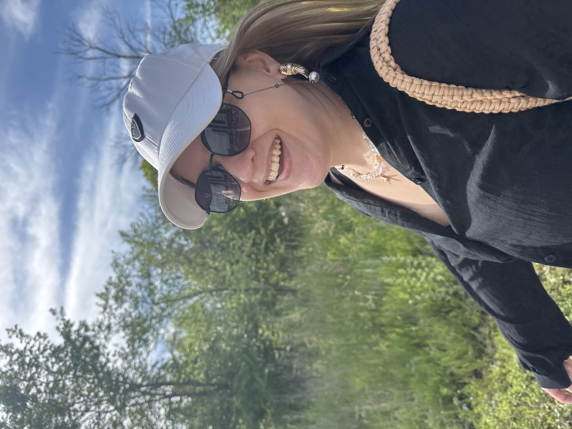 A woman wearing a white baseball cap, sunglasses, and a black button-up shirt smiles as she takes a selfie outdoors. Her hair, a shade of blonde, is visible around the cap. She wears a decorative necklace composed of pearls and other beads, as well as a chain attached to her sunglasses. A textured, woven strap is visible across her shoulder. The background consists of green foliage and a blue sky with wispy white clouds, suggesting daytime and pleasant weather. The image is oriented sideways.