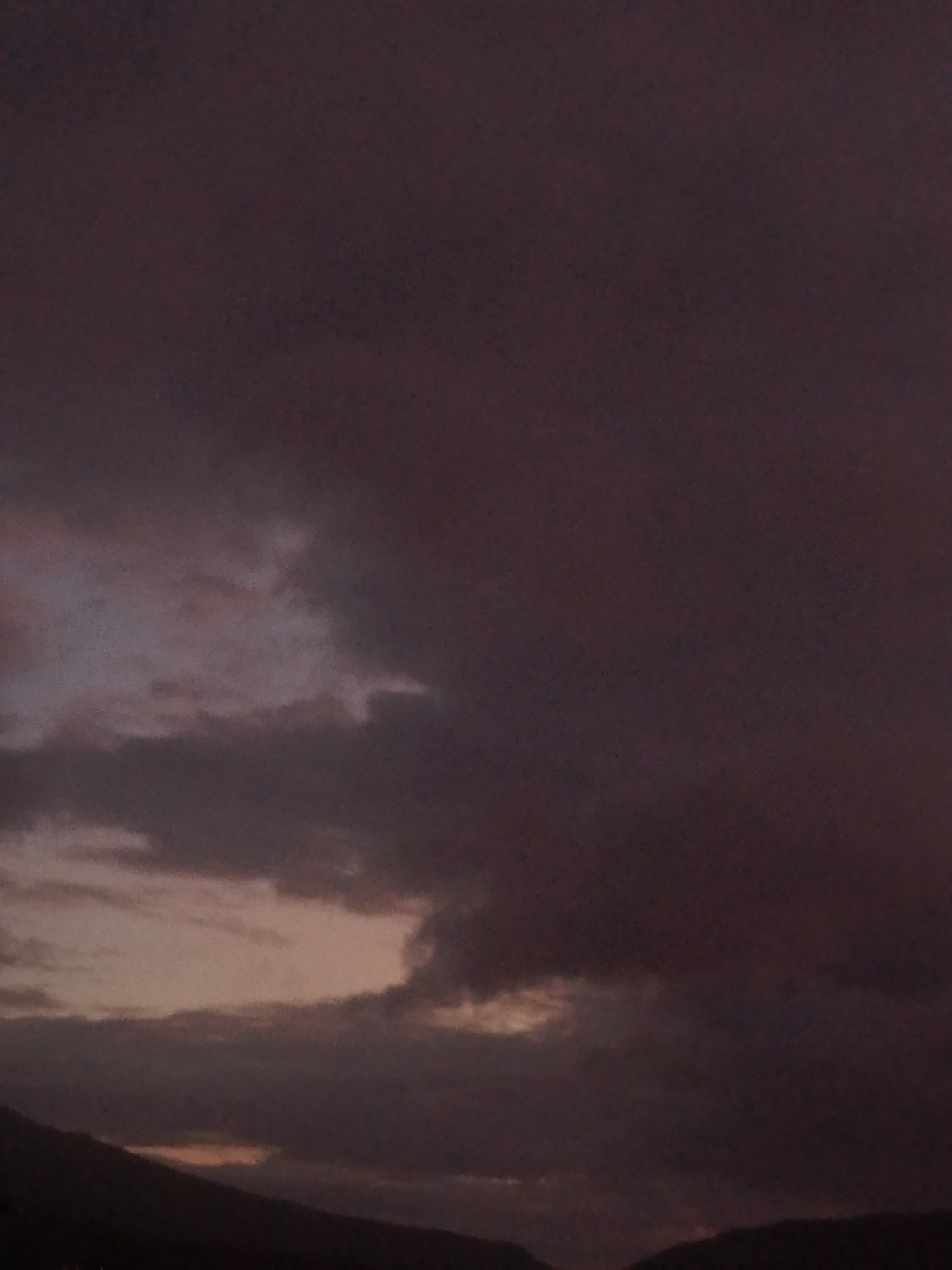 The image shows a dark, cloudy sky, likely at dusk or dawn. The lower portion of the image suggests the presence of land, potentially hills or mountains. The overall tone is somber, with a limited color palette dominated by shades of purple, gray, and dark brown. There are no discernible people, subjects, objects, actions or interactions. There is no way to confirm or deny the location of the image as being in Juan Griego, Venezuela from visual data alone.
