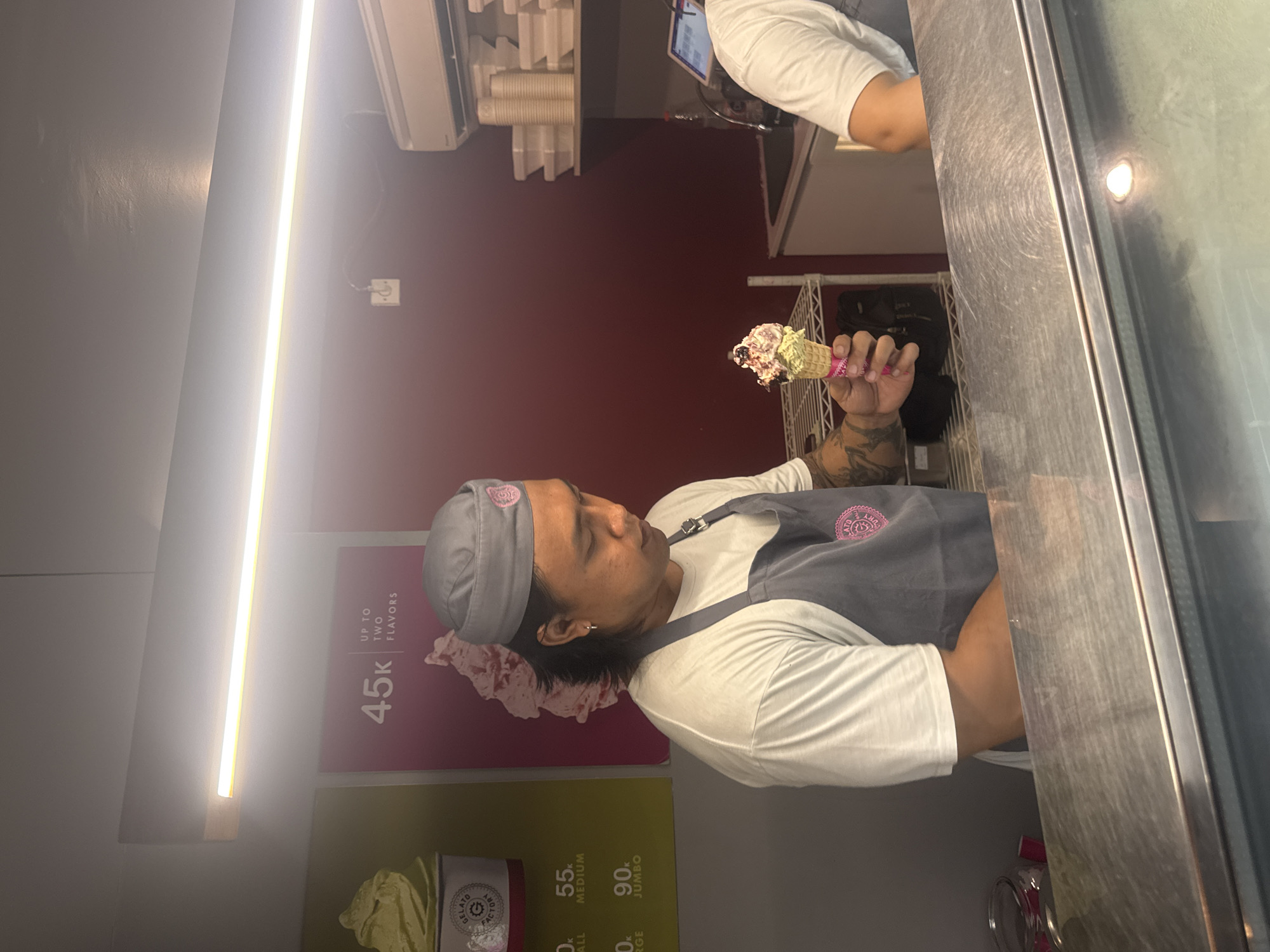 The image depicts an indoor scene at "Gelato Factory," a gelato shop in Kuta Selatan, Indonesia. A male employee wearing a grey gelato hat and apron stands behind a stainless steel counter, holding a cone of gelato. He is looking down at the gelato he is holding. The counter is well-lit by an overhead fluorescent light fixture. Behind the employee, a pink poster displays "45k UP TO TWO FLAVORS" and an image of gelato. To the left of the poster, another poster shows a cup of green gelato. In the background, shelves hold stacks of white takeout containers and cups. Another person, partially visible, stands at the far right of the counter, wearing a white t-shirt. The environment appears clean and organized, typical of a food service establishment.