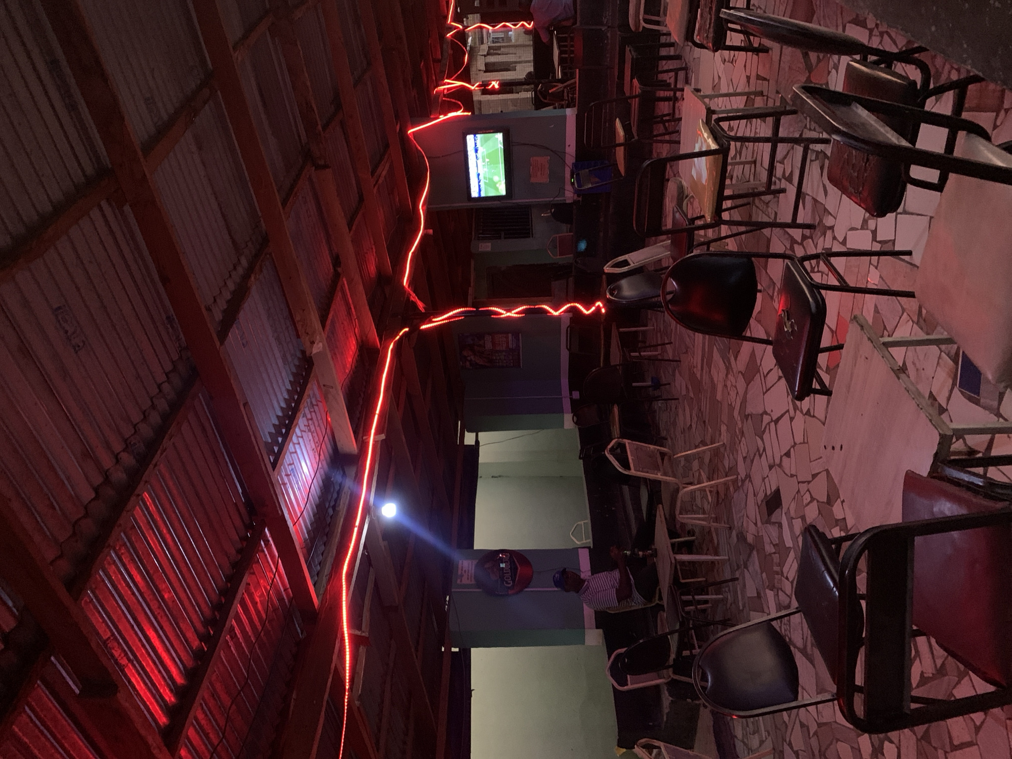 The image depicts the interior of a likely open-air bar in Kabong, Nigeria. The setting is dim, illuminated primarily by a string of red LED lights running along the ceiling beams and a television screen displaying a soccer match. The building has a corrugated metal roof supported by wooden beams. The floor is tiled with a mosaic pattern of various shapes and colors. The space is filled with metal-framed chairs and small tables. Several individuals are seated at the tables, appearing to be patrons. A man is visible in the background wearing a striped shirt and a baseball cap. The bar is decorated with a "GOLD GUINNESS" sign.