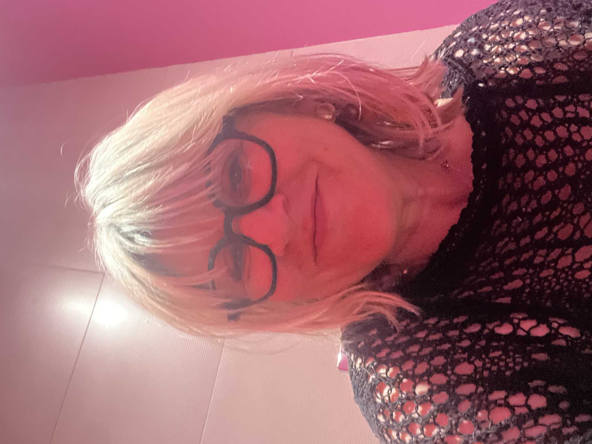 A woman with blond hair and glasses is seen from a low-angle perspective, suggesting she is looking down at the camera. She is wearing a black mesh top. The background is pink and white, possibly a wall. The location is unspecified and there is nothing to suggest it is Montrond-les-Bains, France.