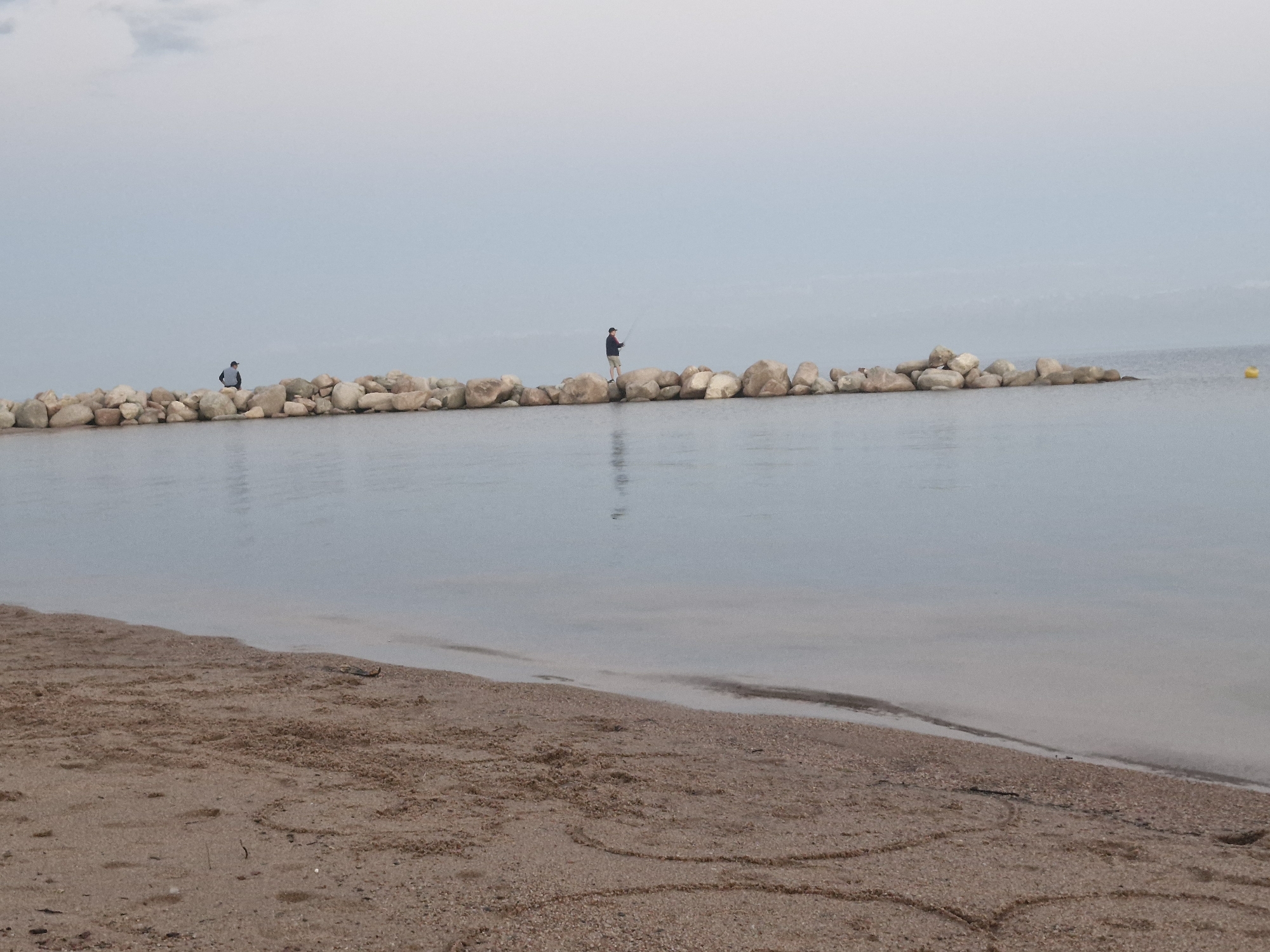 The image depicts a serene, overcast day at a beach, likely in or near Bulan-Segettu, Kyrgyzstan, though the location is not explicitly confirmed within the image itself. In the distance, a line of large, light-colored rocks forms a breakwater or pier extending into the calm, gray water. Two individuals are visible on this rocky structure. One person, further to the left, is standing and appears to be observing the water. The second person, positioned more centrally, is actively fishing with a rod. The water is still, reflecting the muted tones of the sky and the rocks. The foreground consists of a sandy beach, appearing damp near the waterline, with some subtle patterns etched into the sand, possibly from the tide or wind. A small yellow buoy is visible in the far right of the water. The overall atmosphere is tranquil and subdued.