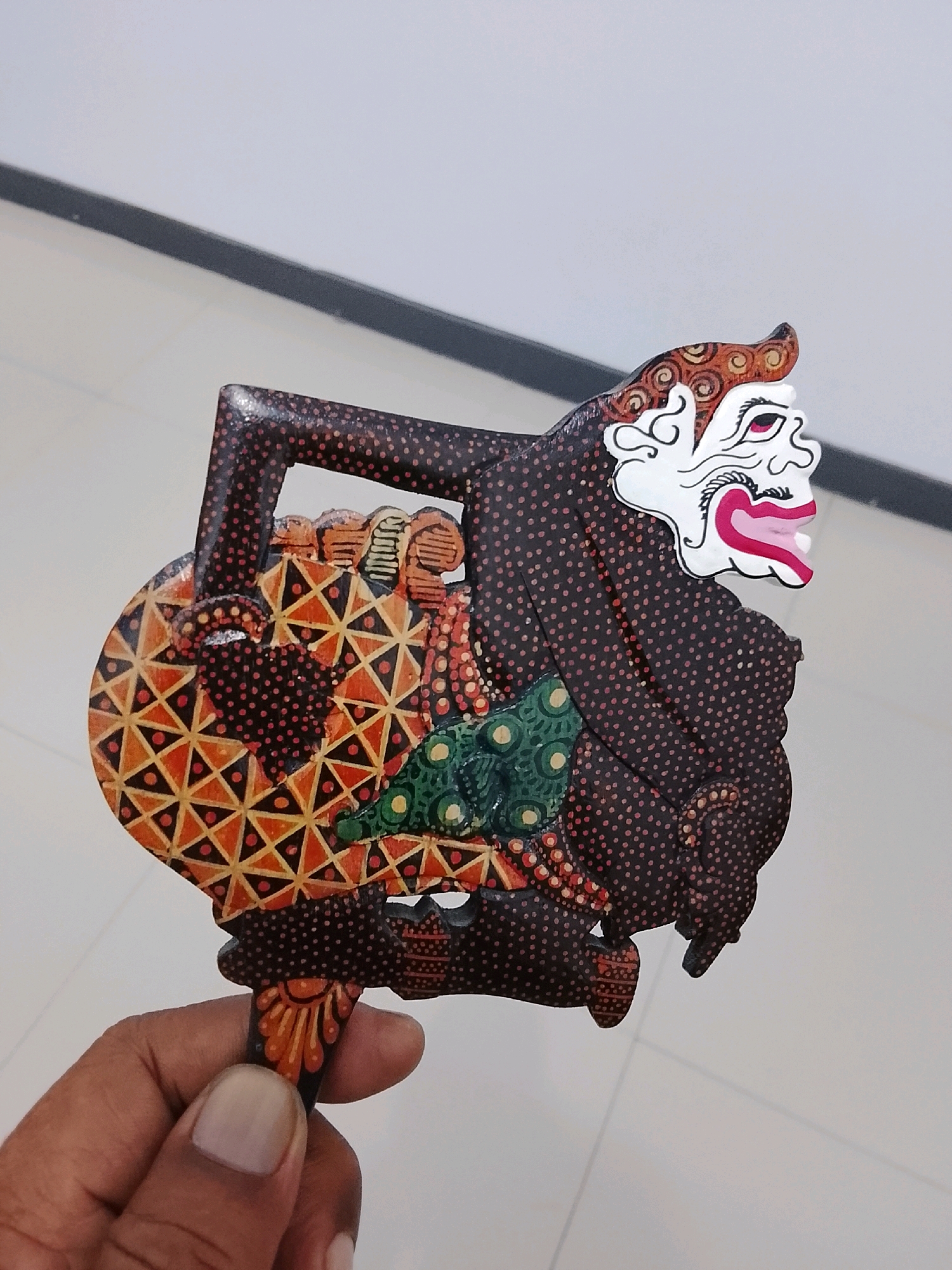 An overhead shot captures a hand holding a traditional Javanese wayang golek puppet. The puppet is crafted from wood and painted with intricate details. It features a dark brown body adorned with small red dots, representing clothing. A white face with expressive red eyes and pink lips stands out, framed by a stylized brown and orange headdress. The puppet's arms and legs are visible, along with a circular shield on its left side, decorated with geometric patterns of orange, yellow, and red triangles on a black background. A green patterned fabric is visible on the puppet's torso. The background consists of a light-colored floor with subtle linear patterns, possibly tiles, and a dark horizontal line running across the upper portion of the image, suggesting a wall or partition. The scene appears to be indoors, with the lighting casting soft shadows. The location is identified as Slawi, Indonesia, but no specific landmarks or environmental features are discernible in the image to confirm this.