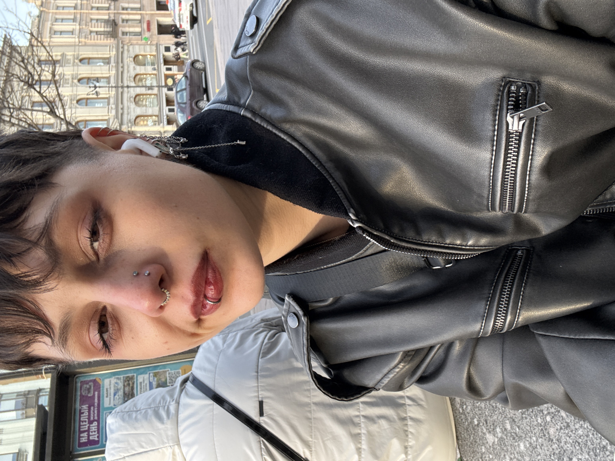 A person is centered in the frame, wearing a black leather jacket, and has multiple piercings in their nose and lip. They are wearing an earbud, with a chain earring attached. The background shows a city street in Saint Petersburg, Russia, with buildings and parked cars visible. A bus stop sign is also visible to the left of the person. Another person, dressed in a white puffer jacket with a black strap across their chest, is partially visible in the lower part of the frame. The image appears to have been taken at a slight angle, possibly by the person in the leather jacket.