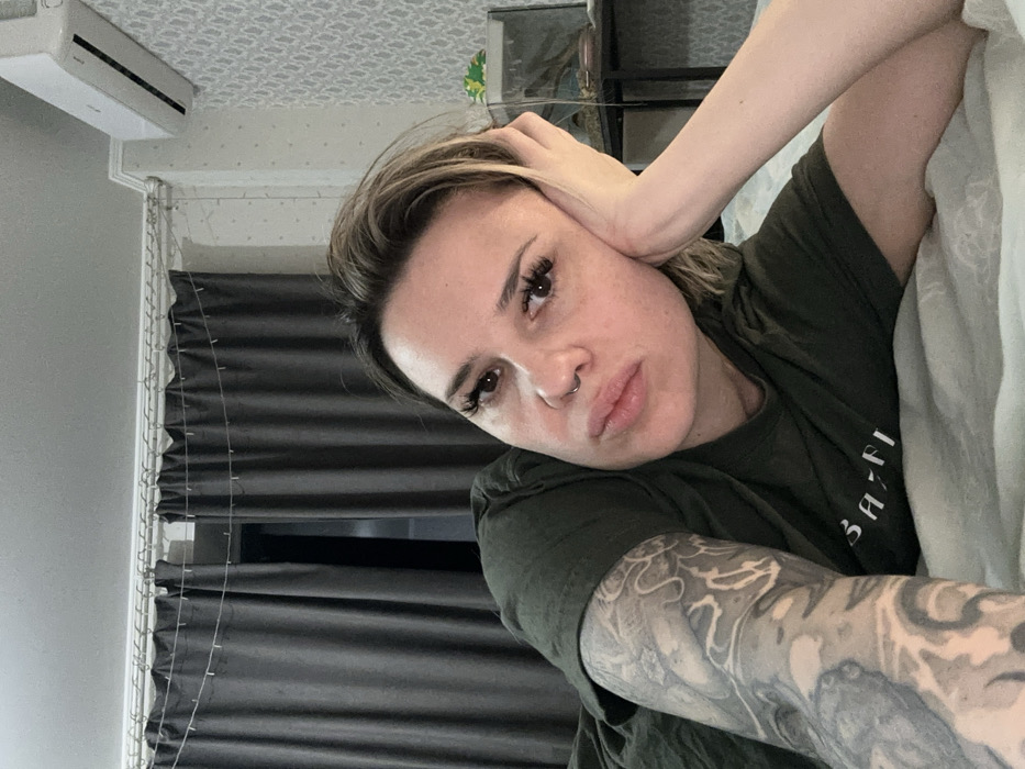 A female subject is depicted in a medium close-up, lying on a light-colored bed or bedding. She has light-colored hair with darker roots, prominent false eyelashes, and a silver nose ring on her left nostril. Her lips are slightly pursed, and her expression is neutral. She wears a dark olive green short-sleeved t-shirt, revealing extensive grey-toned tattoos covering her visible left arm. Her right arm is bent, with her right hand supporting the right side of her head.
The background shows dark grey curtains covering a window, across which a string of small, unlit decorative lights is suspended. To the right, a light grey patterned wallpaper covers the wall. A white wall-mounted split-system air conditioning unit is installed near the ceiling in the upper right quadrant of the image. Further to the right, a clear glass enclosure, possibly a terrarium or aquarium, rests on a dark shelf, containing substrate and some green elements. The scene is an indoor residential setting in Rostov-on-Don, Russia.